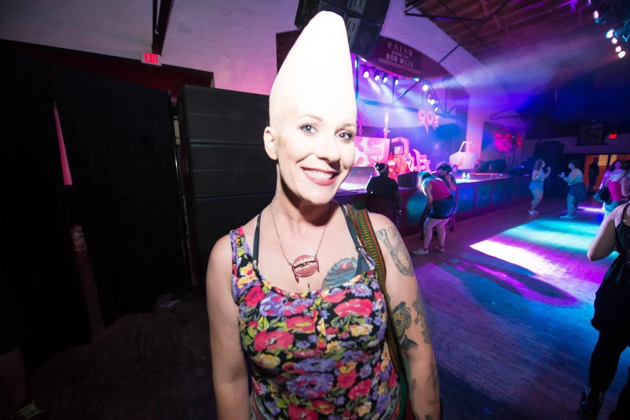A woman with a tall, pointed hairstyle and tattoos, smiling at a party with colorful lighting, other people, and a decorated stage in the background.
