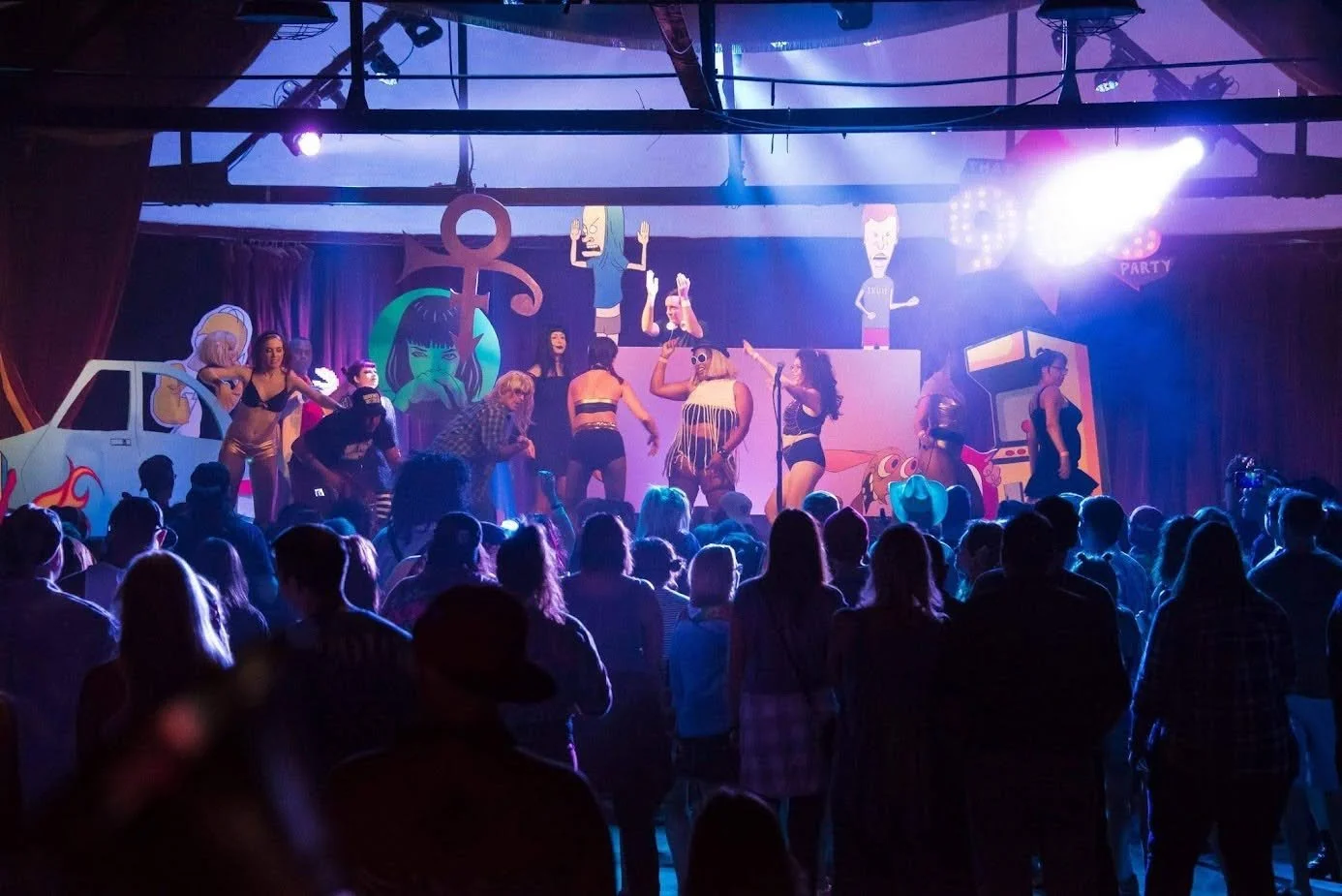 A lively stage performance with dancers in colorful costumes, animated cartoon-themed decorations including a large anchor, a car, and cartoon characters, with an enthusiastic crowd watching in a dimly lit venue.