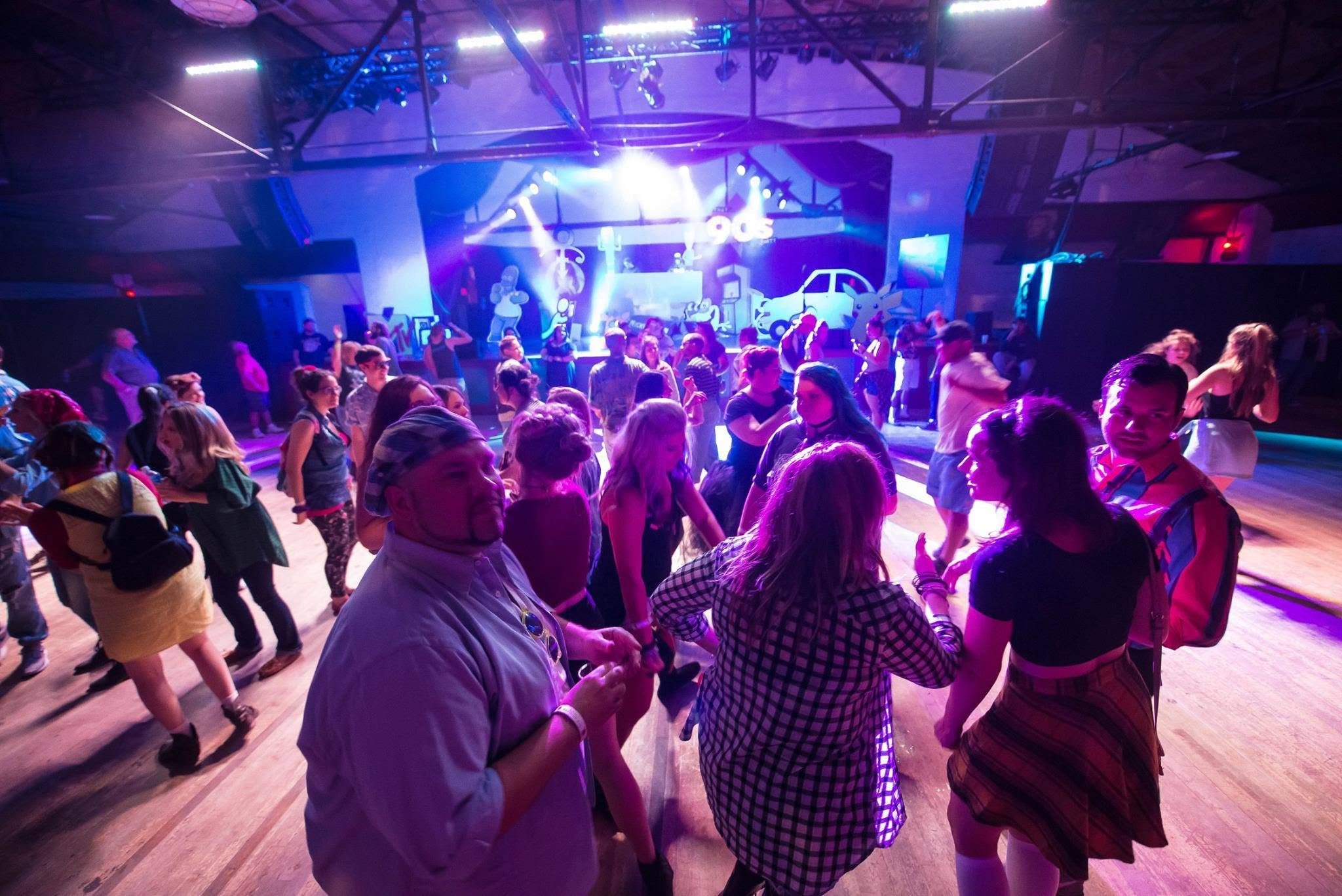 People dancing at a party with colorful lights and a DJ on stage.