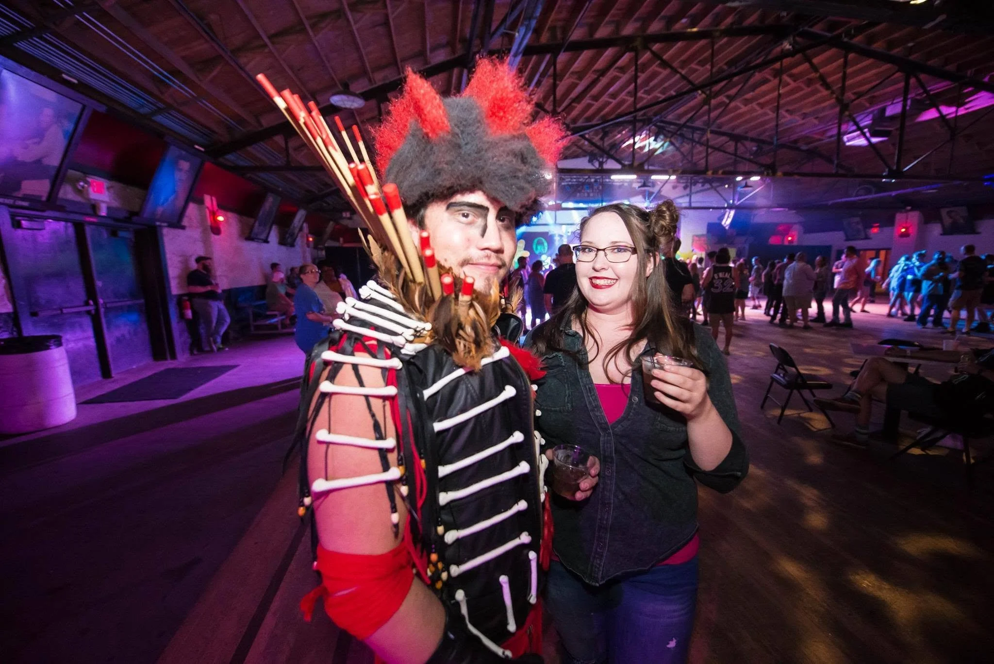 A man with face makeup and a costume with bones and feathers, standing beside a woman with glasses and long hair, holding drinks at a lively indoor event with colorful lighting, dancing, and people in the background.