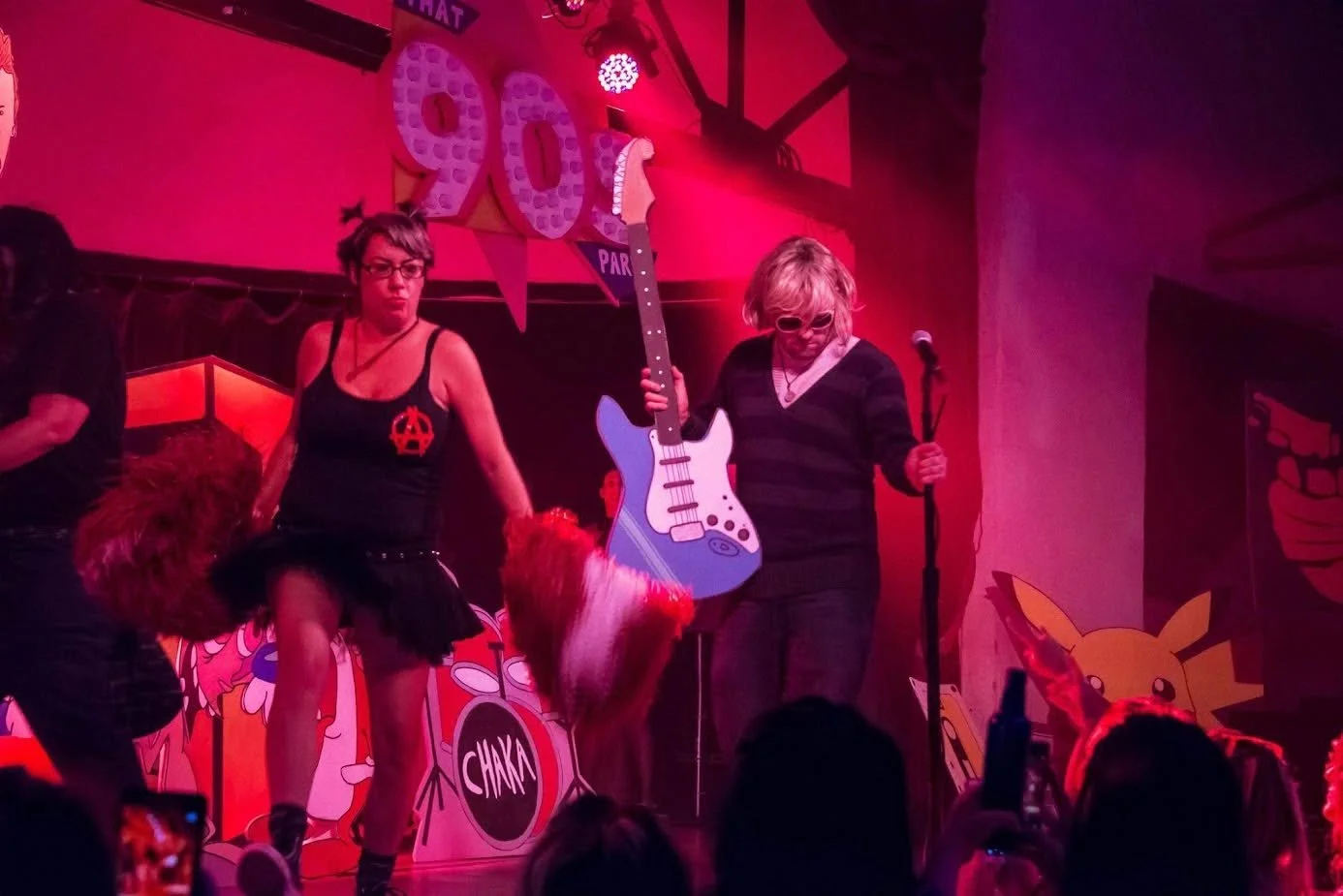 People on stage at a Pokémon-themed event, with a woman in a black tank top and a tutu, another woman holding a giant plastic guitar, and Pikachu illustrations in the background.