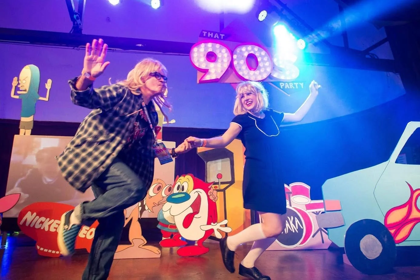 Two women dancing and holding hands at a 90s themed party with colorful cartoon characters and signs in the background.