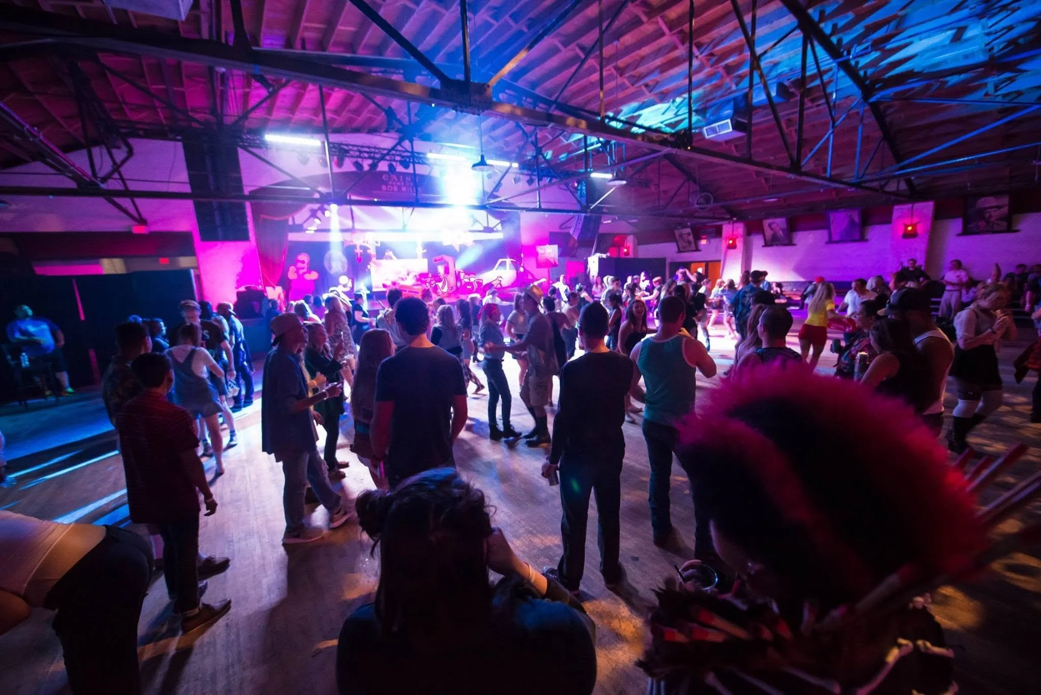 People dancing in a large indoor venue with colorful stage lighting and framed pictures on the walls.
