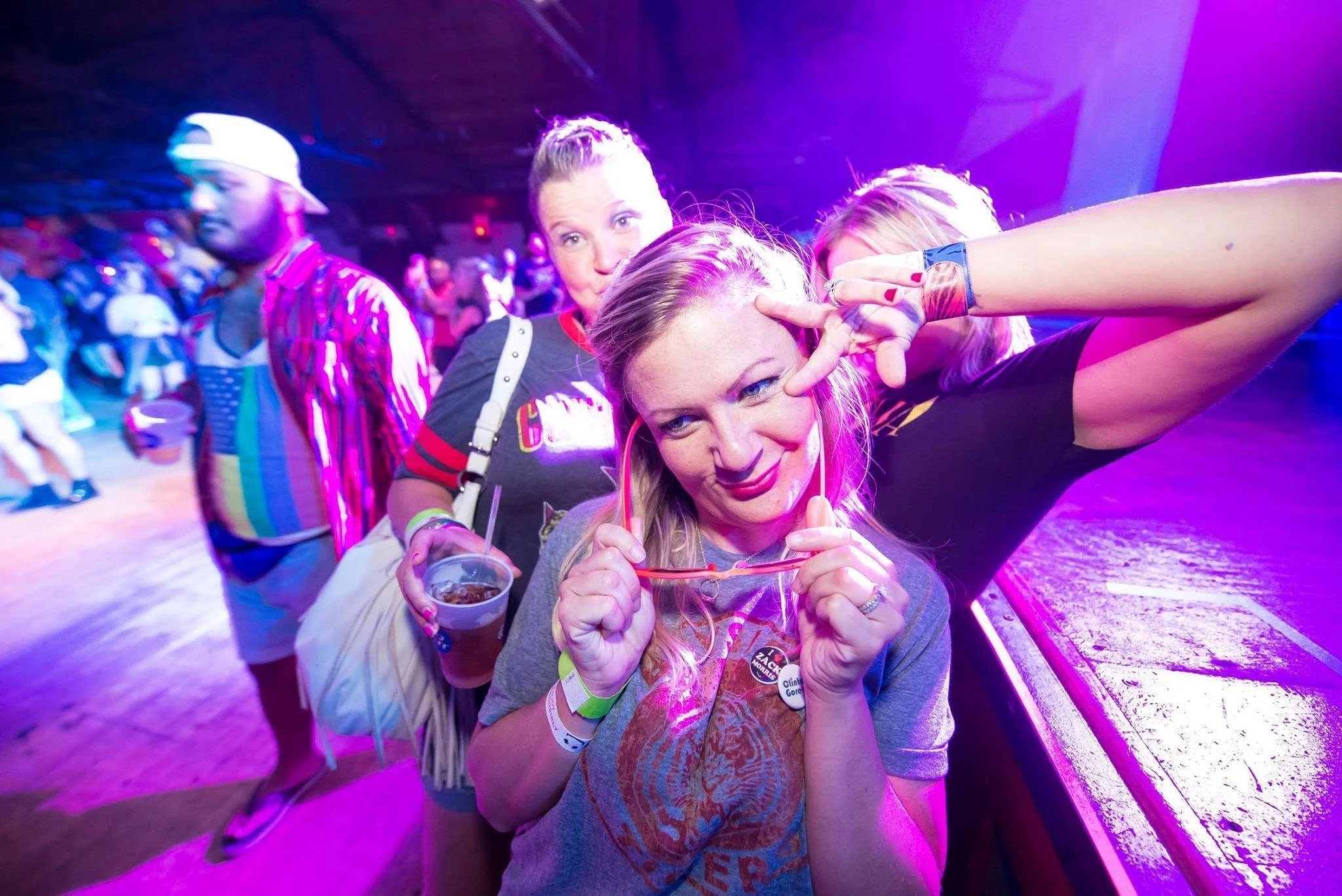 A group of women enjoying themselves at a party or concert, with purple lighting, one making a peace sign, holding a drink and wearing a button, others in colorful attire and wristbands.