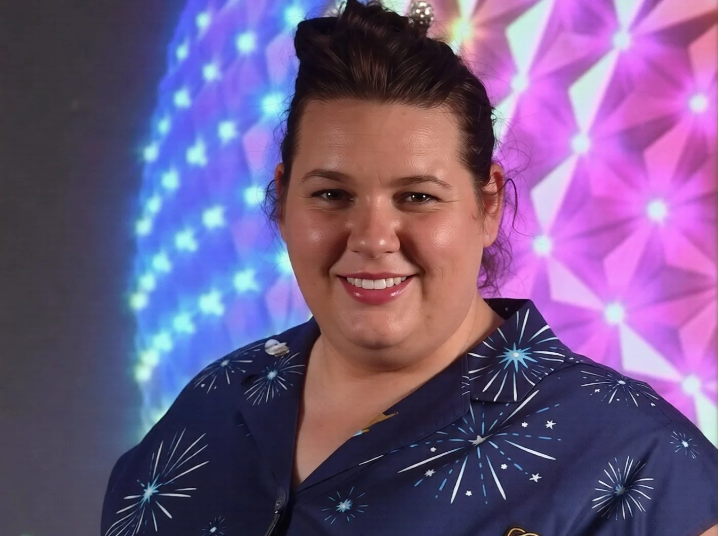 A person with smiling face, dark hair styled upwards, wearing a shirt with firework patterns, stands in front of a colorful background of illuminated fireworks.