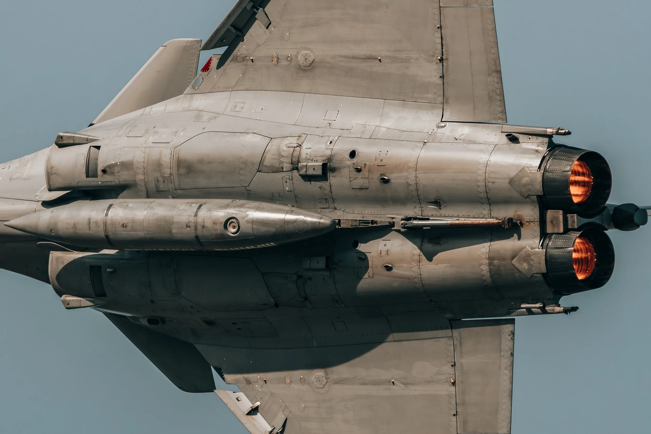 Fuselage d'un avion de chasse en vol, montrant les moteurs en arrière avec des flammes sortant, en arrière-plan un ciel clair.