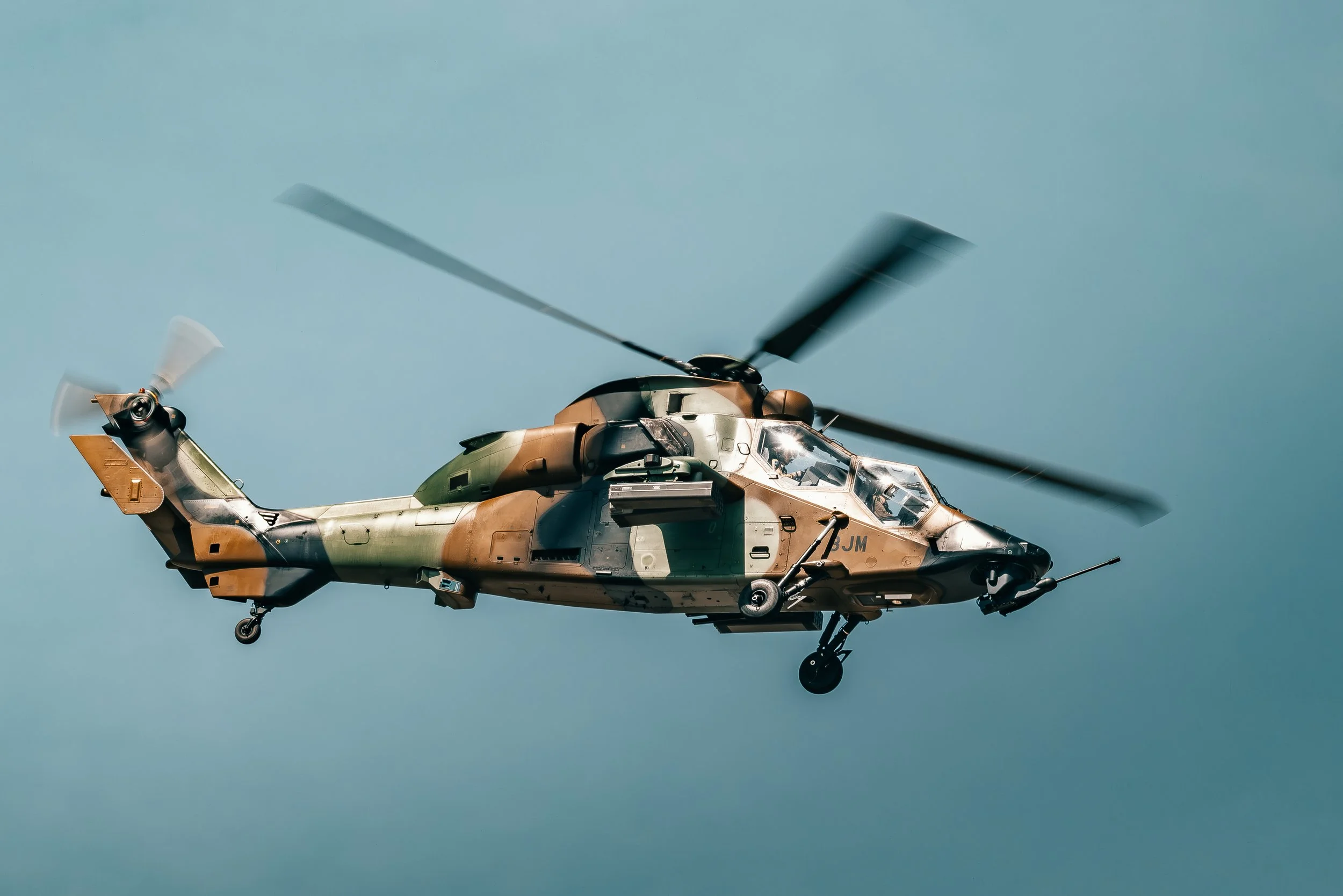 Un hélicoptère de combat en vol, camouflage vert, marron et noir, contre un ciel bleu clair.