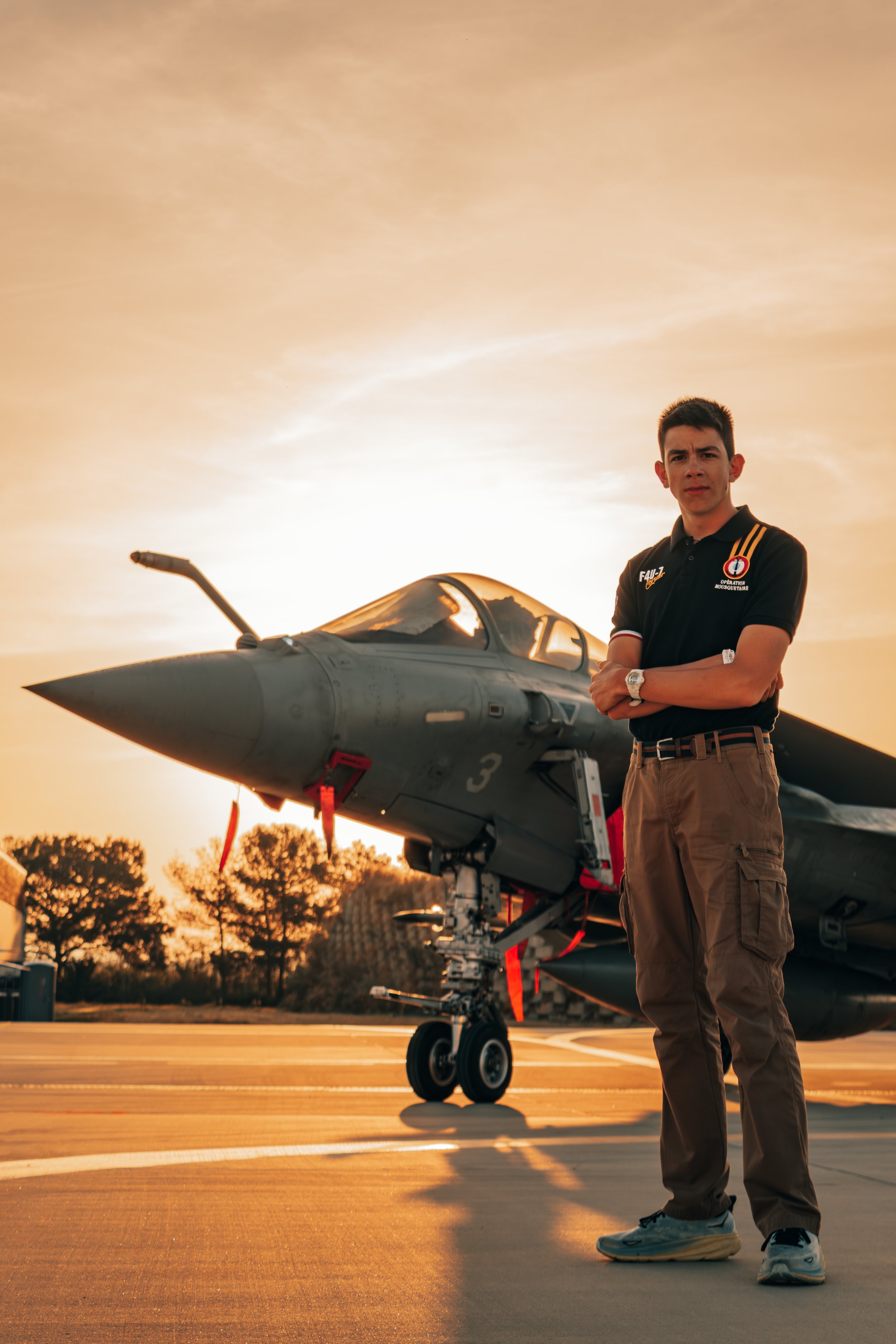 Un jeune homme debout devant un avion de chasse sur un tarmac lors d'un coucher de soleil, portant une chemise noire avec des logos, pantalon cargo beige et baskets, avec les bras croisés.