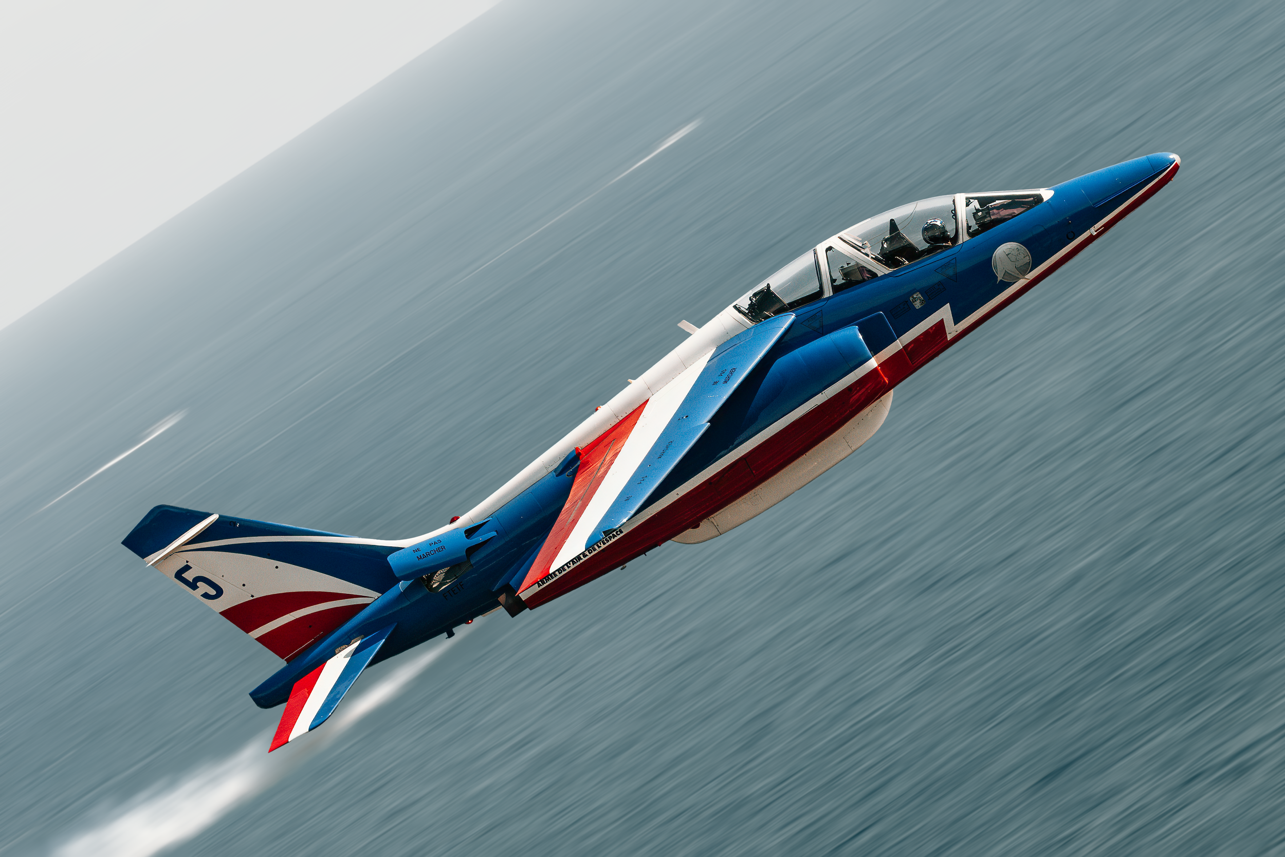 Un avion de chasse de couleur bleue, blanche, rouge, volant à grande vitesse dans le ciel, laissant une traînée derrière lui.