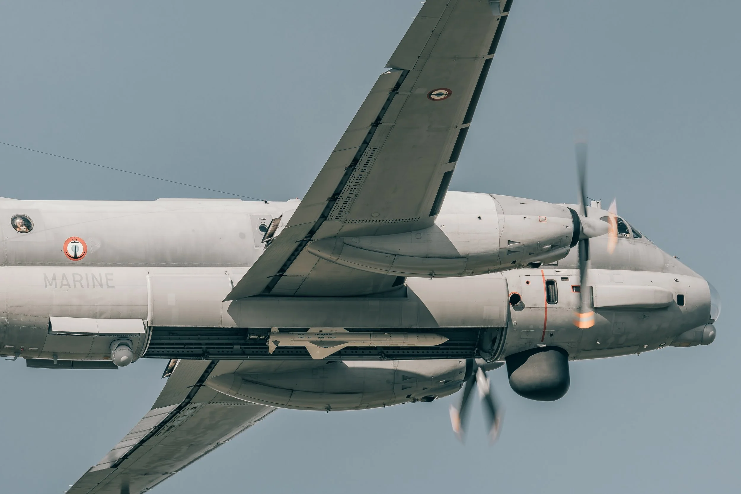 Un avion militaire blanc vu de côté avec deux moteurs et une hélice en marche, fond de ciel bleu, marquages militaires.