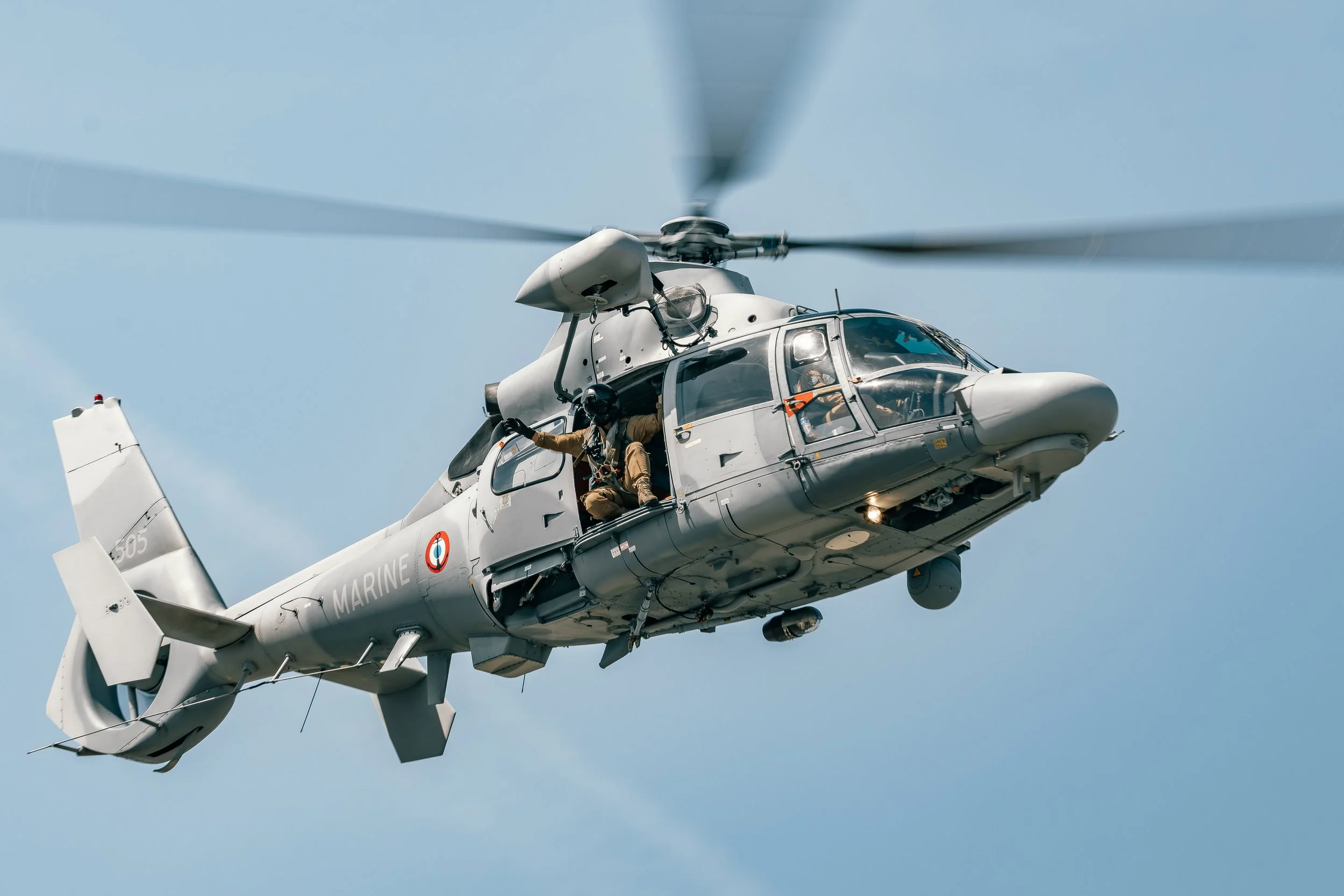 Un hélicoptère de la marine française en vol, avec un marin à l'ouverture de la porte, portant un casque et un uniforme, dans un ciel clair.