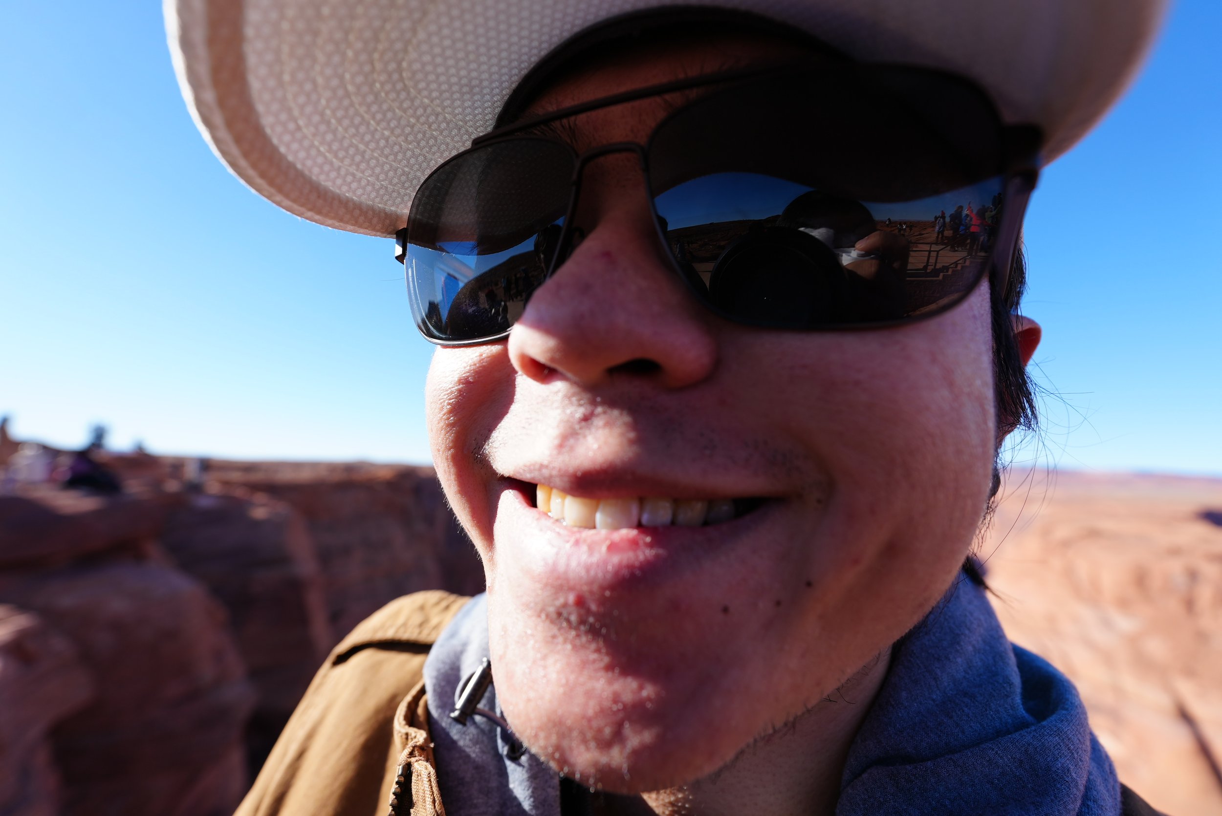 Close-up of a person wearing a hat and sunglasses, smiling outdoors with a clear blue sky in the background.