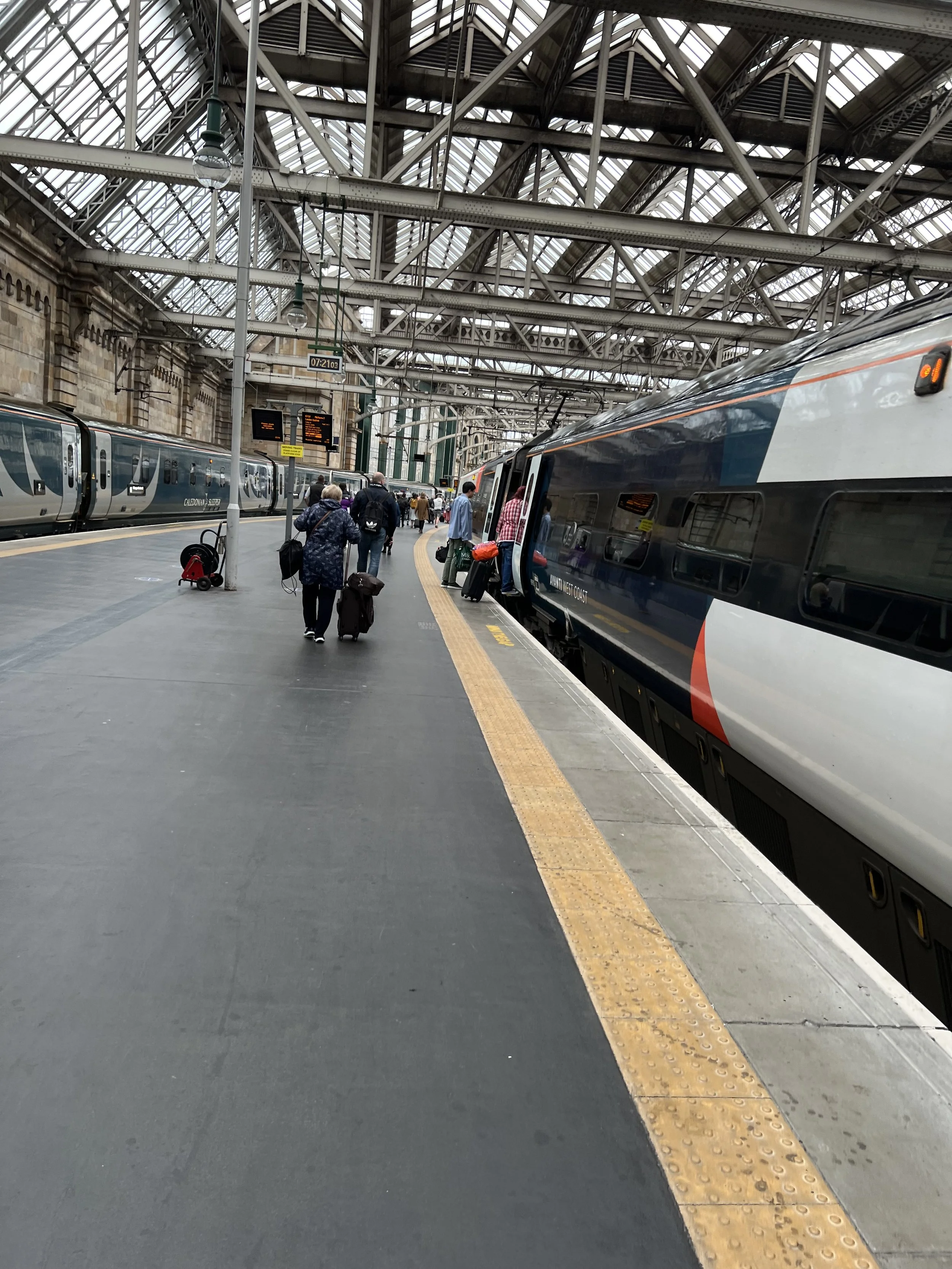 People boarding and leaving Caledonian Sleeper trains at a busy train station with a glass and steel roof structure.