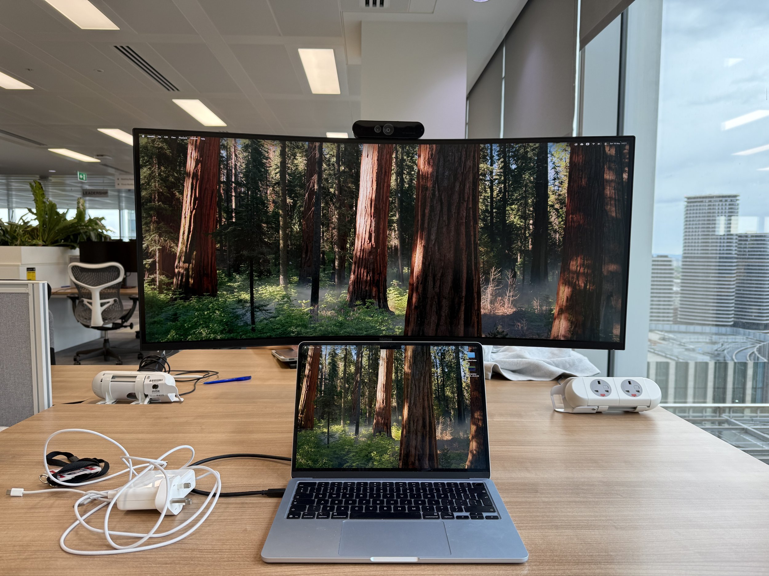A desk setup with a laptop, a monitor displaying a forest scene of trees and greenery, a smartphone, power strips, and tangled chargers. Background shows office chairs, potted plant, large windows, and an urban cityscape with skyscrapers.