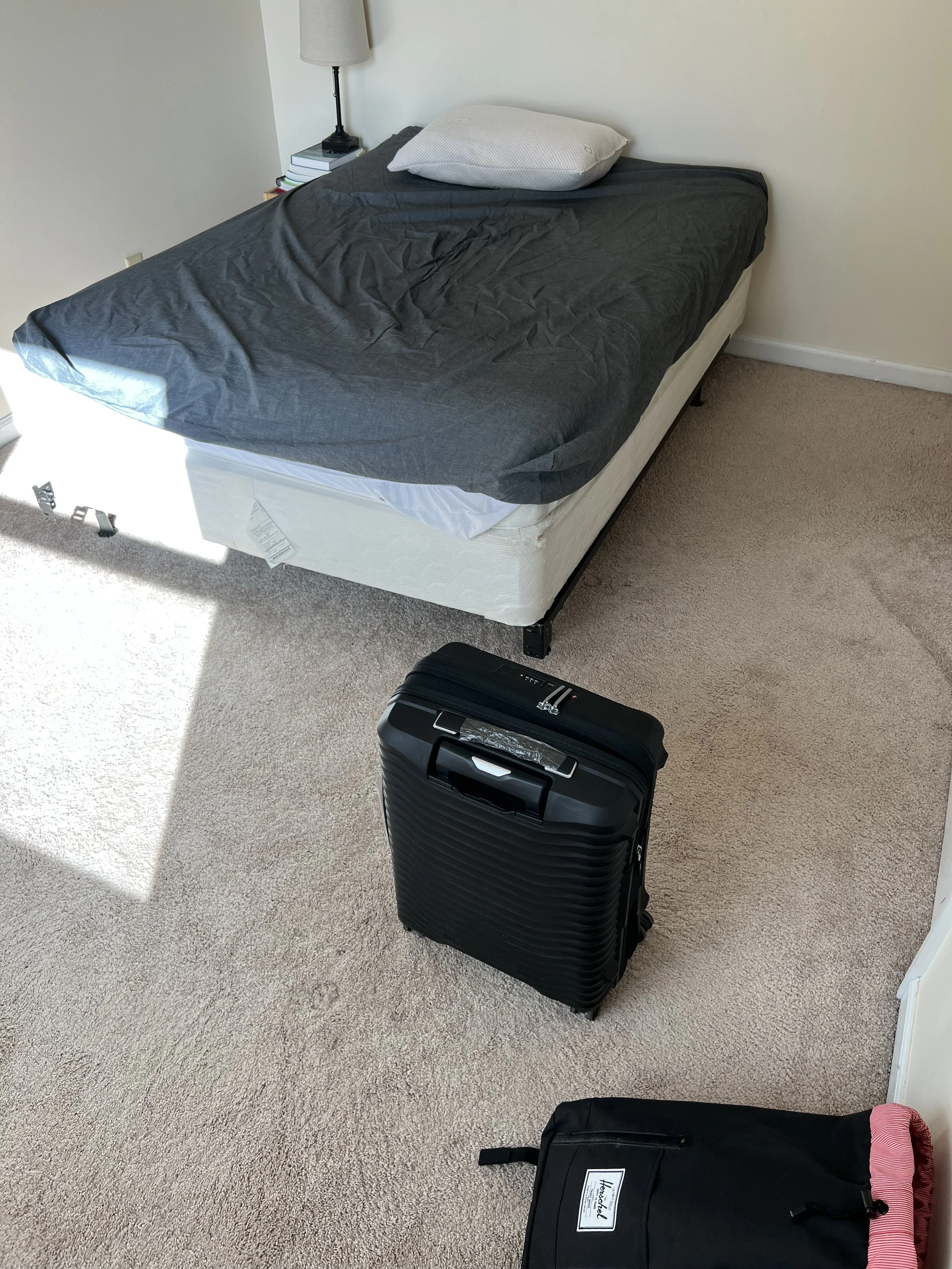 Hotel room with a bed, nightstand, bedside lamp, a small stack of books, a suitcase, and a black bag.
