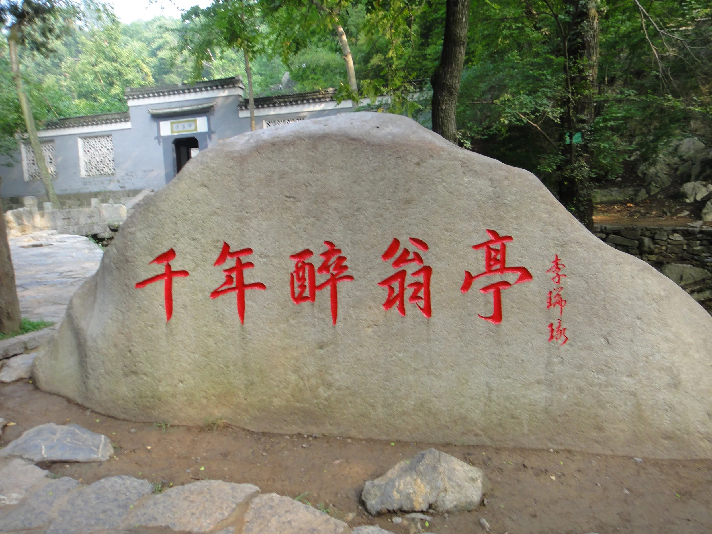 A large gray rock with red Chinese characters carved into it, situated in a forested area with trees and a small traditional structure in the background.