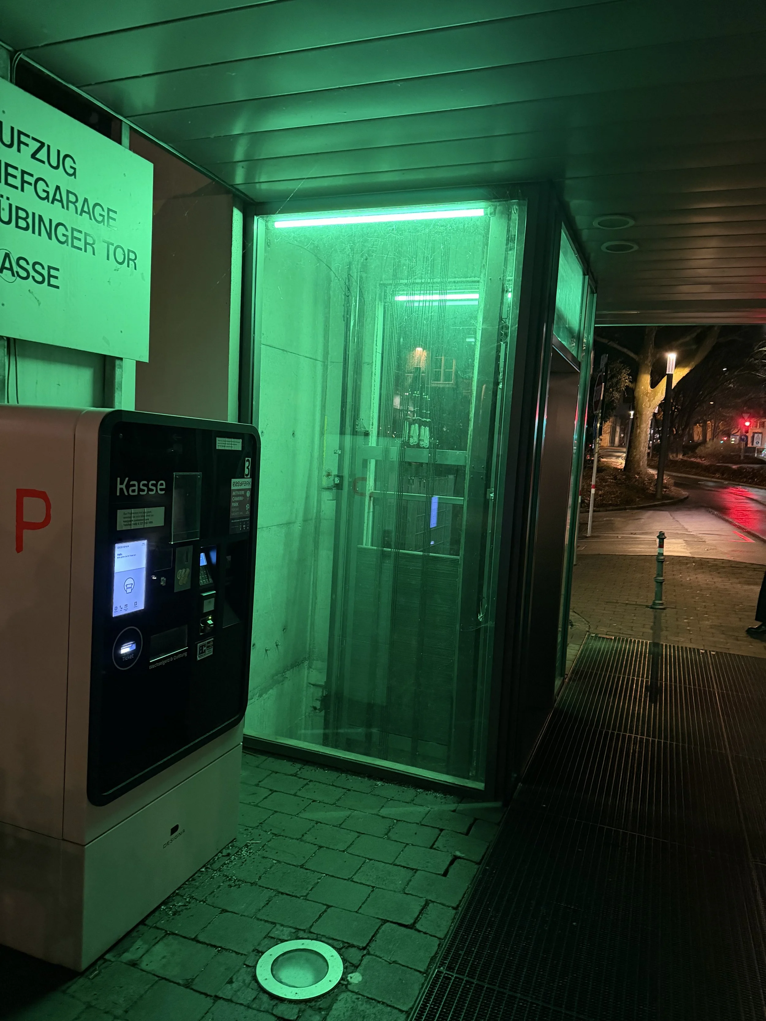 Night scene of a green-lit glass bus stop shelter with an electronic ticket vending machine beside it. The sidewalk is wet, and street lights and trees are visible in the background.