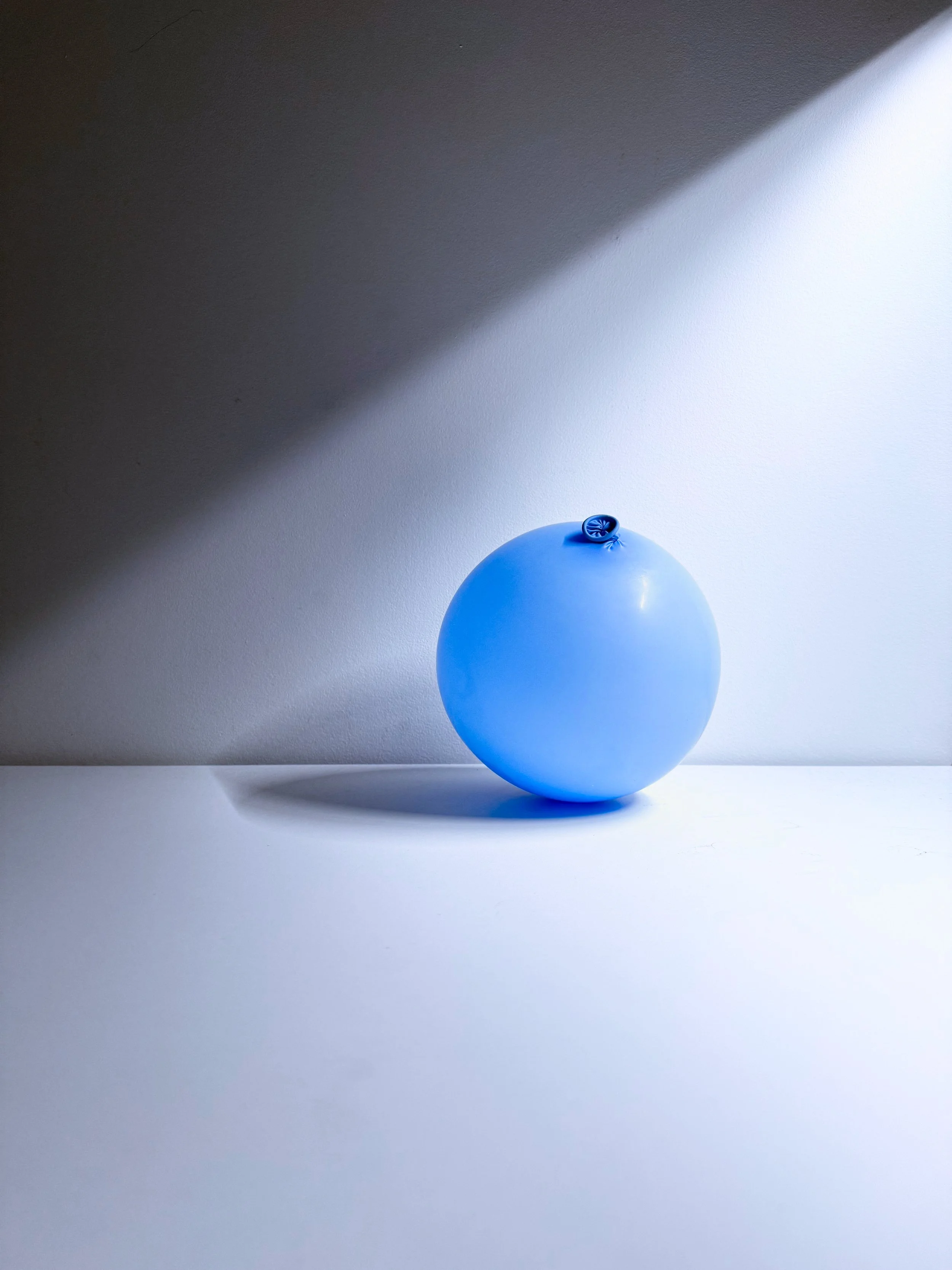 A blue balloon sitting on a flat white surface against a light background, with shadows cast on the wall behind.