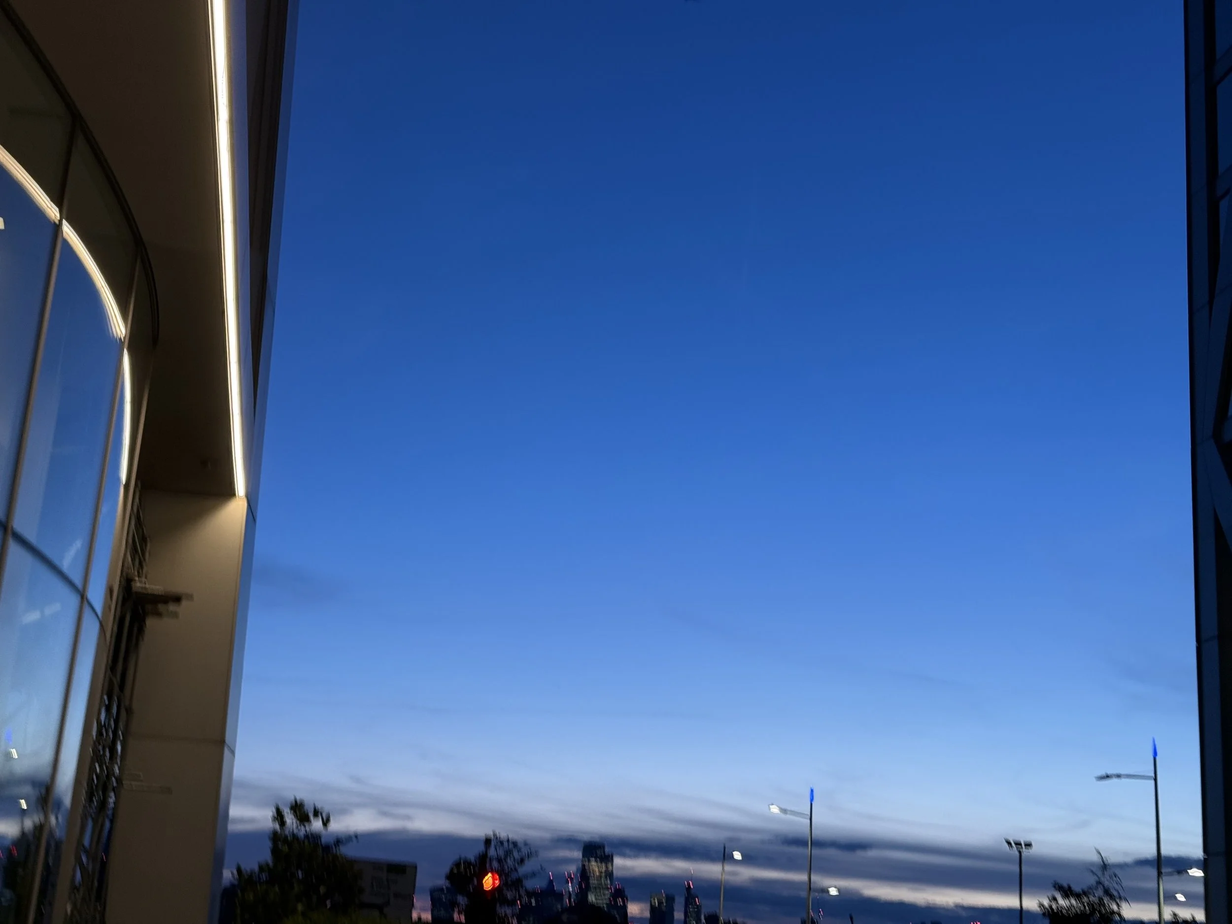 View of the sky during sunset with a dark blue horizon, showing two buildings on the sides and trees below, with streetlights and cars visible.
