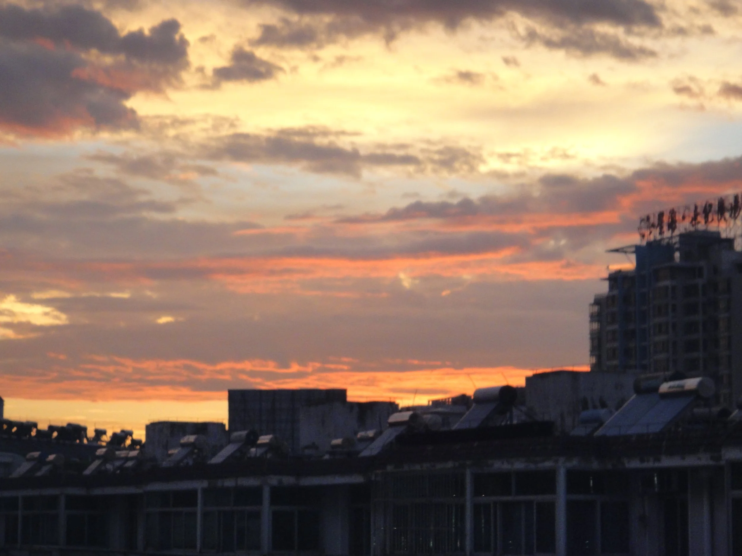 A sunset sky with clouds in shades of orange, yellow, and gray over rooftops and buildings in an urban area.