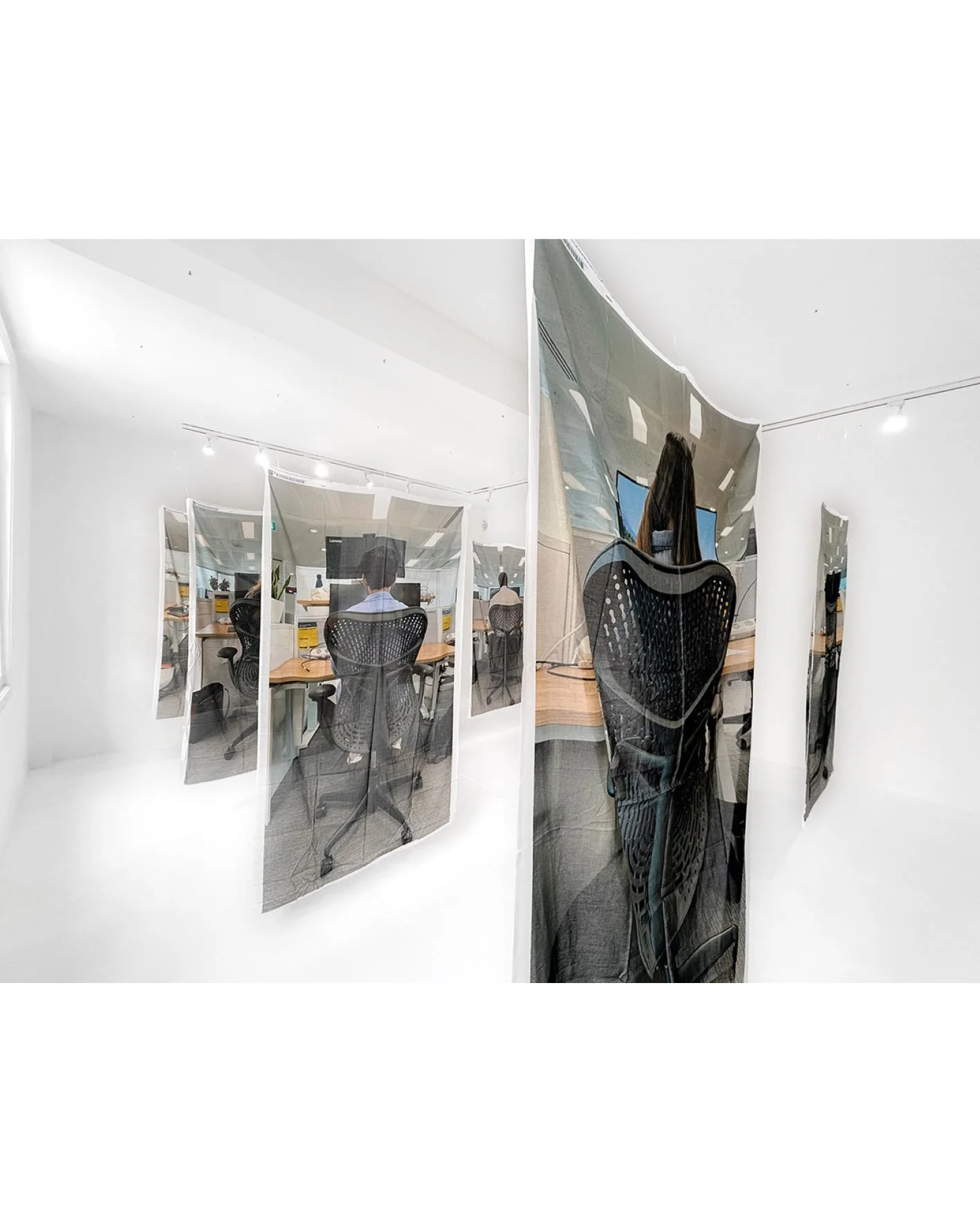 Art installation of multiple mirrored fabric panels in white-walled gallery, reflecting people working at desks in a modern office.