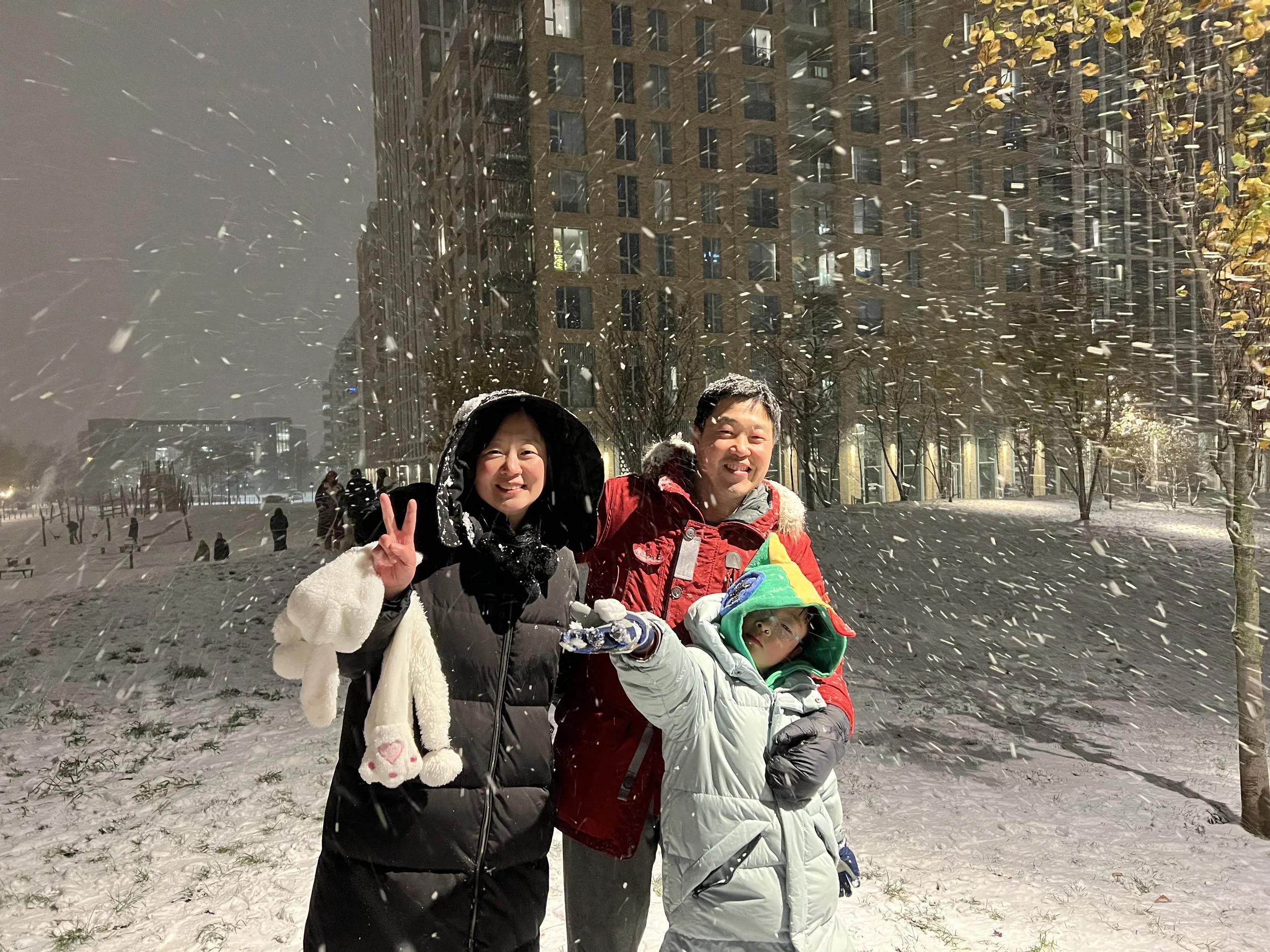 Two smiling people, a woman and a man, standing in a snowy city park at night. The woman is holding a peace sign and wearing a black jacket with a white furry scarf. The man is wearing a red and gray winter coat and holding a child in a white snowsui