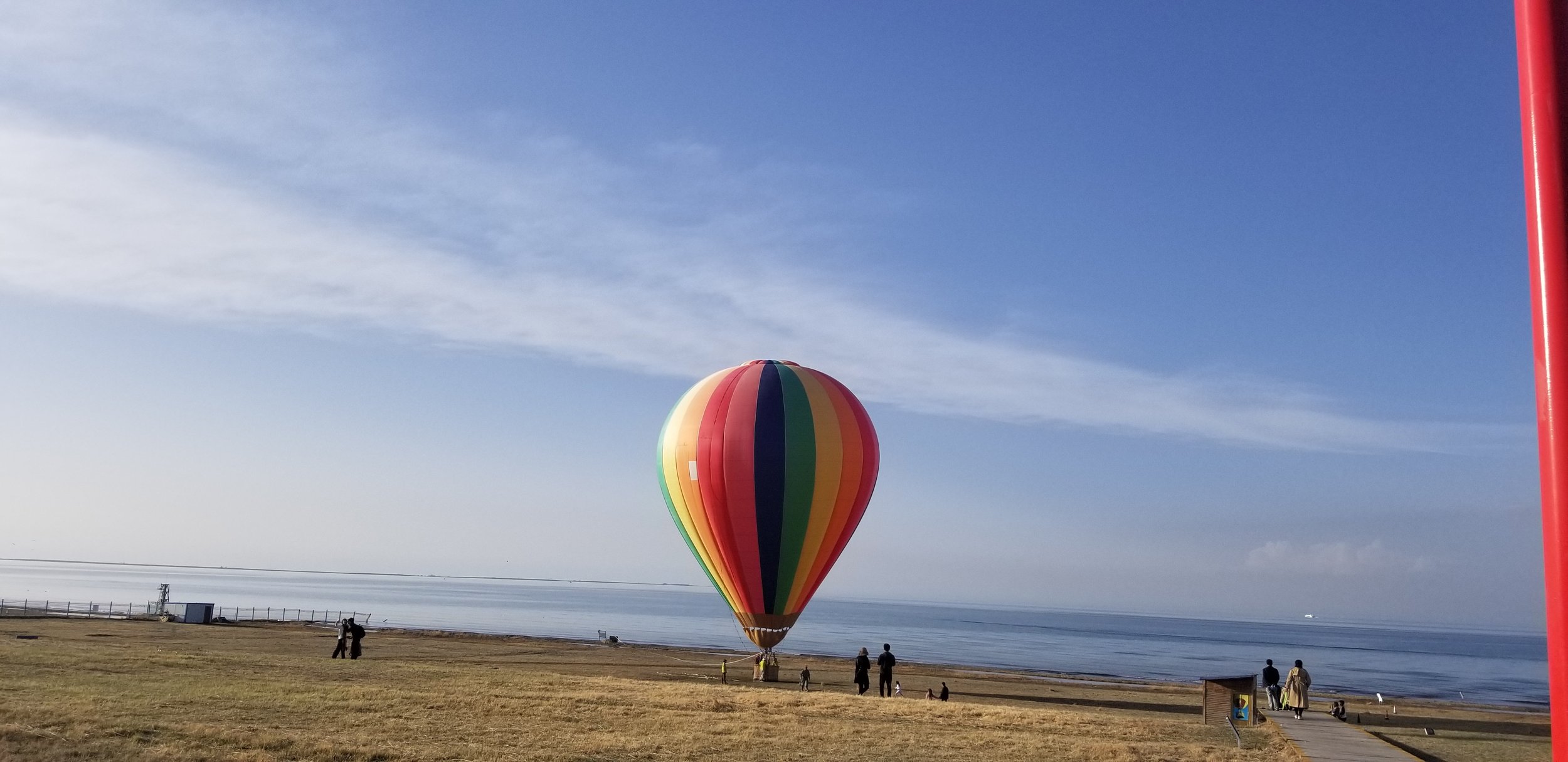 Colorful hot air balloon flying over a grassy park near the water with people walking and a clear blue sky.