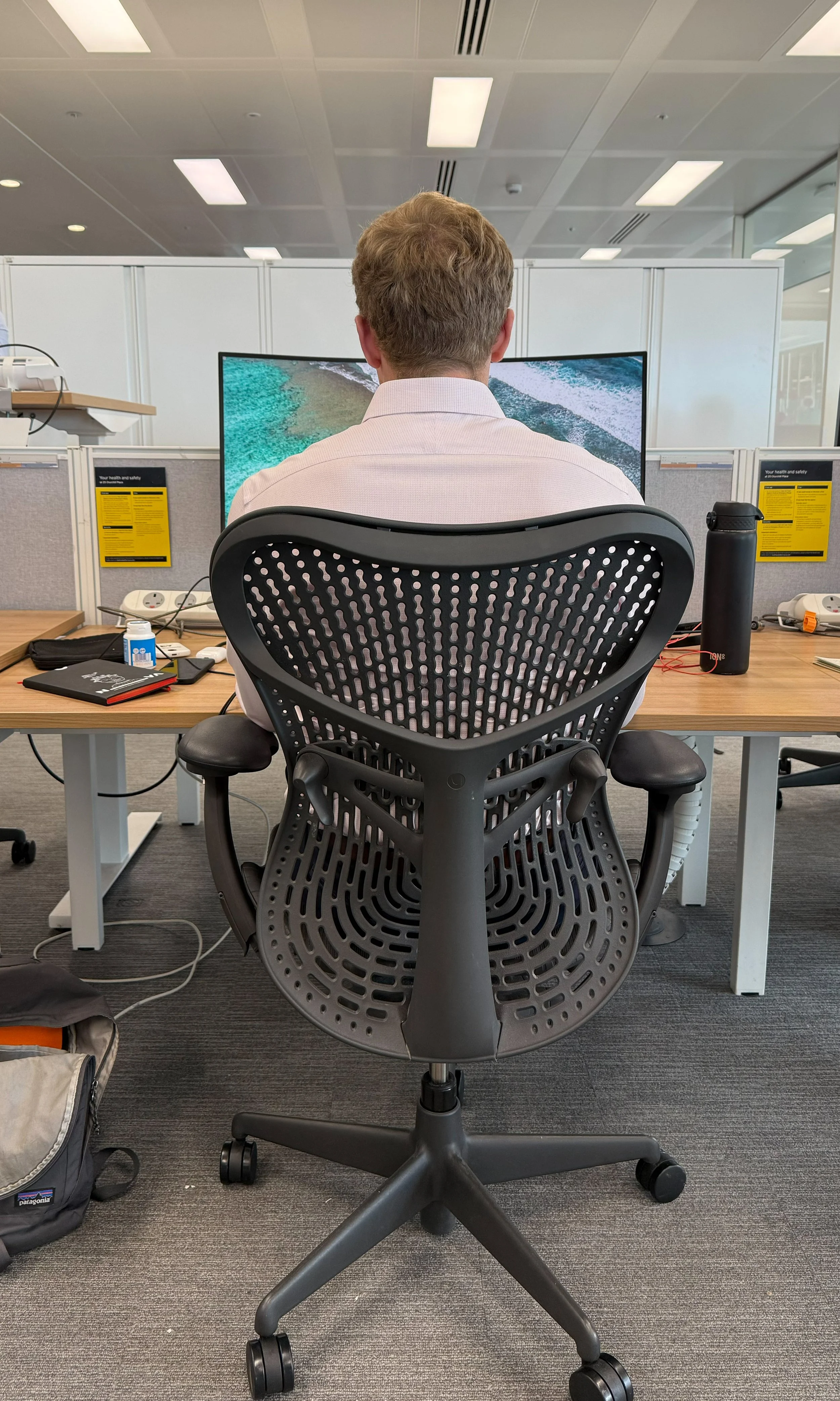 Back of a man sitting at an office desk in front of a curved computer monitor displaying a beach scene. The desk has various items, including a notebook, a water bottle, and some gadgets. The office environment has cubicles, ceiling lights, and equip