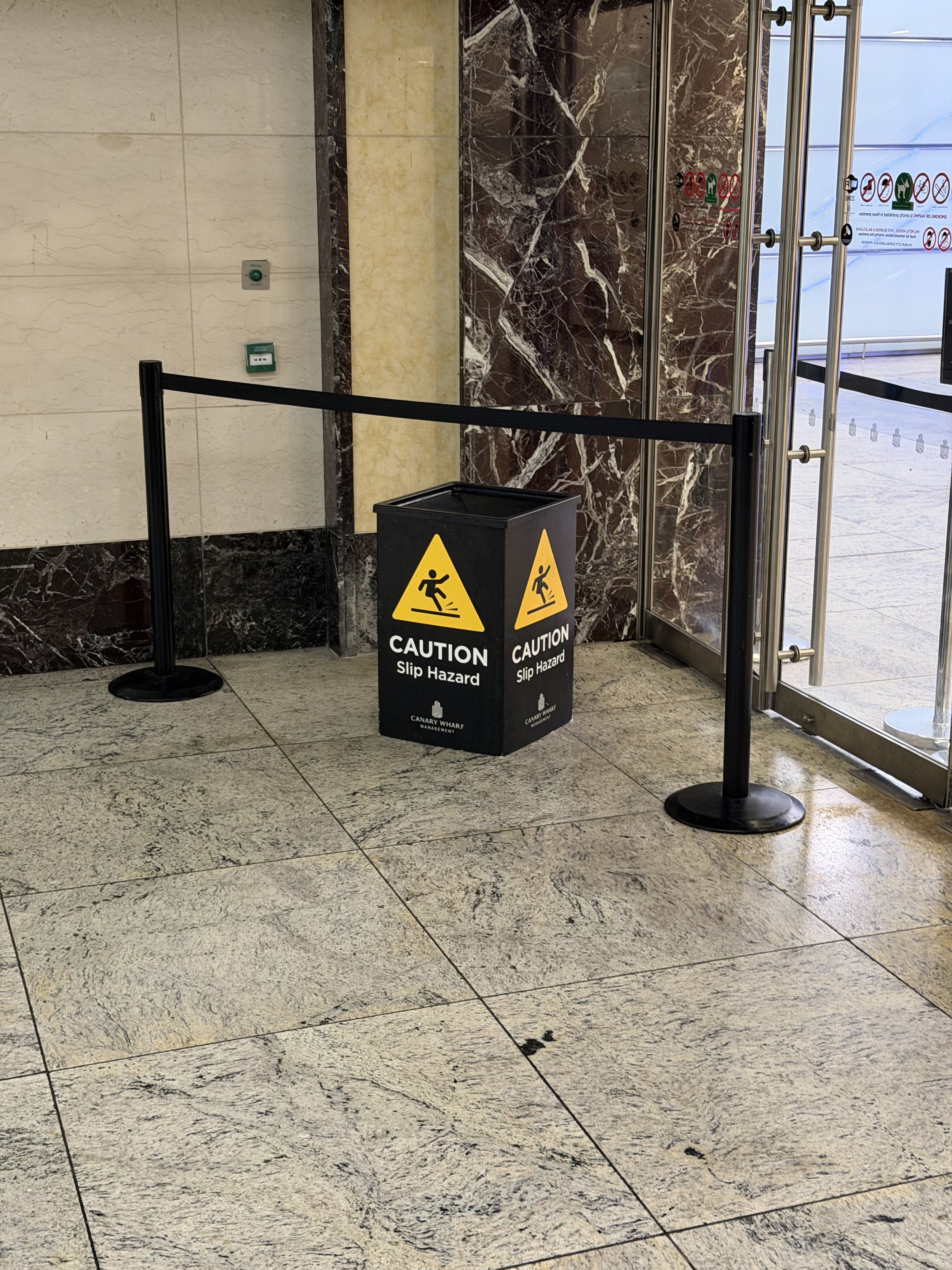 Interior of a building with tiled marble walls and floors, a black caution slip hazard sign, and a door with safety instructions.