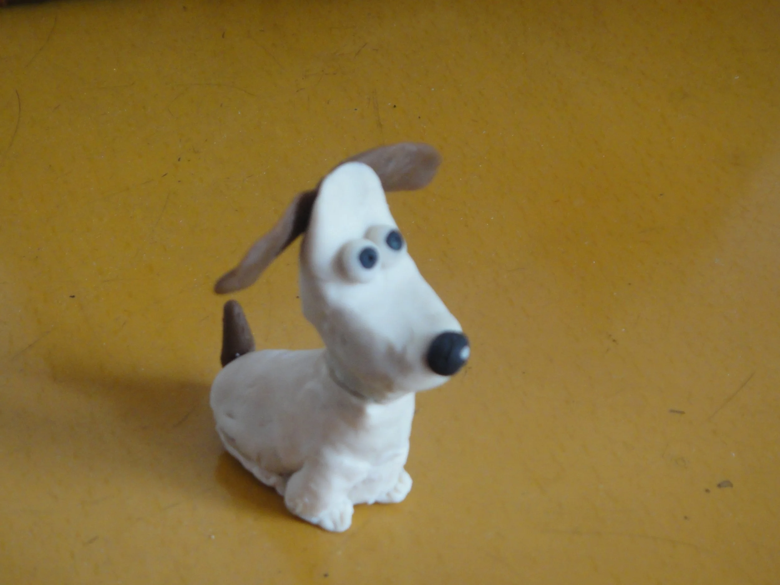 A small sculpture of a dog with blue eyes, a white body, and brown ears and tail, placed on a yellow surface.