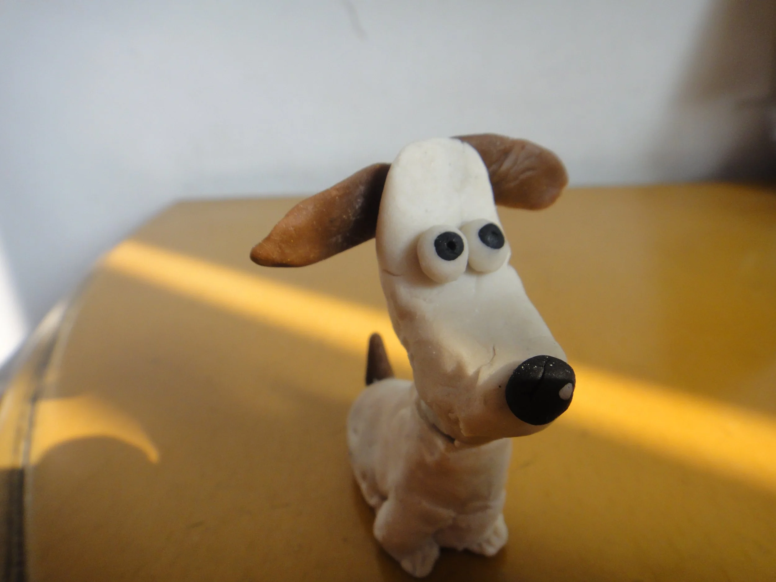 A small craft figure of a dog made from white, brown, and black modeling material with large eyes and floppy ears, standing on a yellow surface.