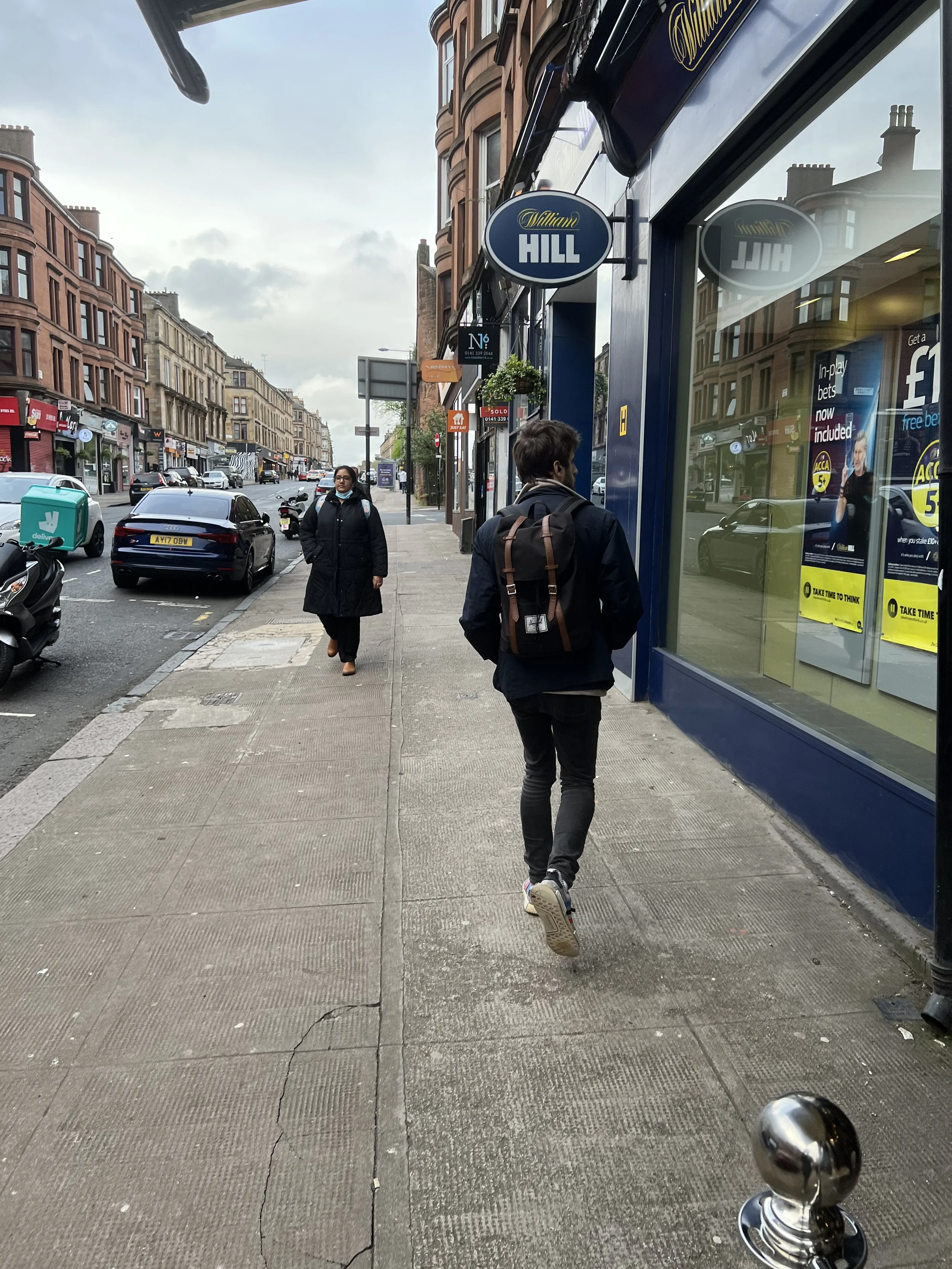 City sidewalk scene with two pedestrians, one man with a backpack walking away and a woman wearing glasses and a coat facing forward, storefronts, parked cars, motorcycles, and a cloudy sky in the background.