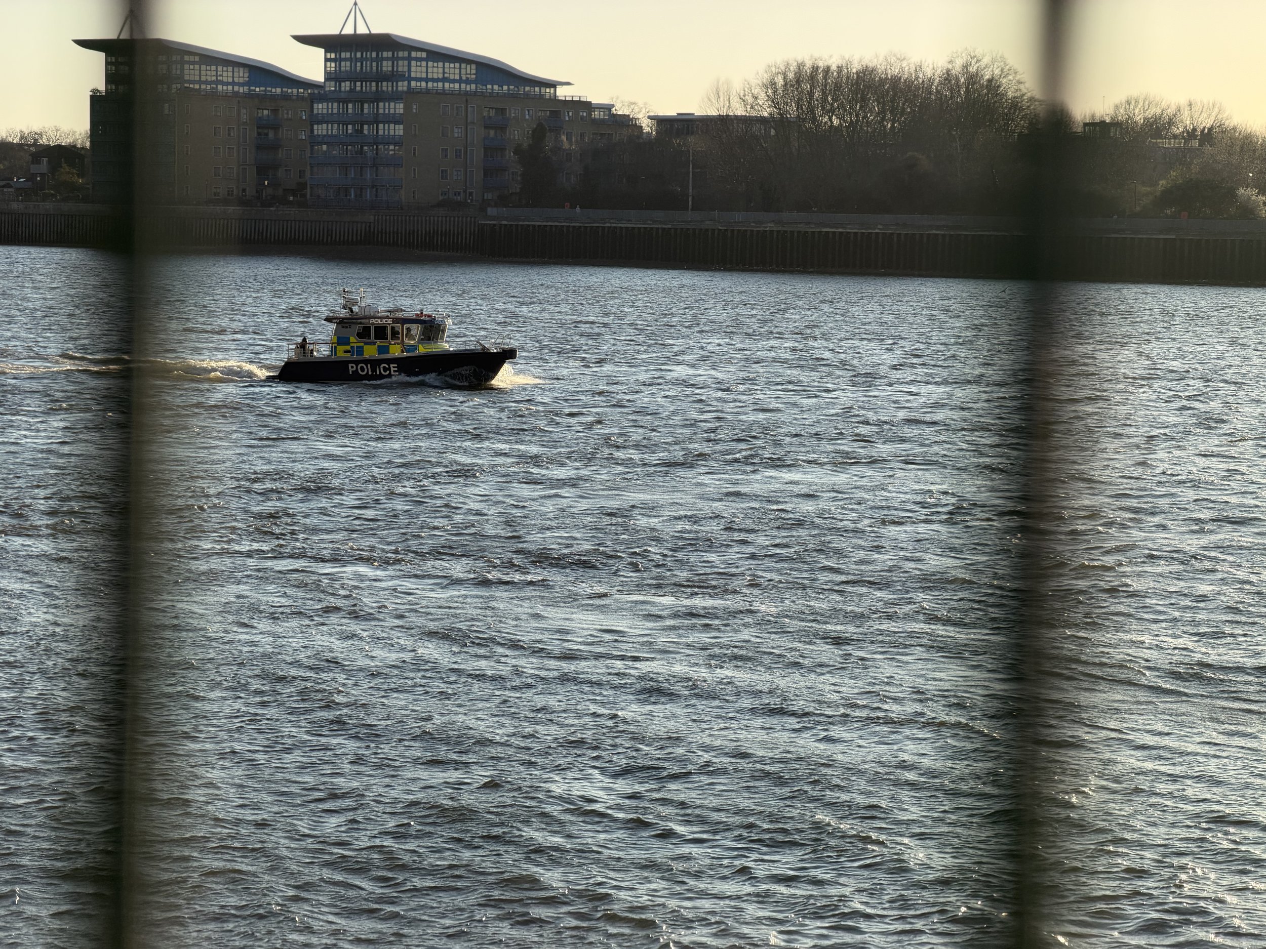 A police boat on a river, with modern residential buildings and trees in the background, seen through a fence.
