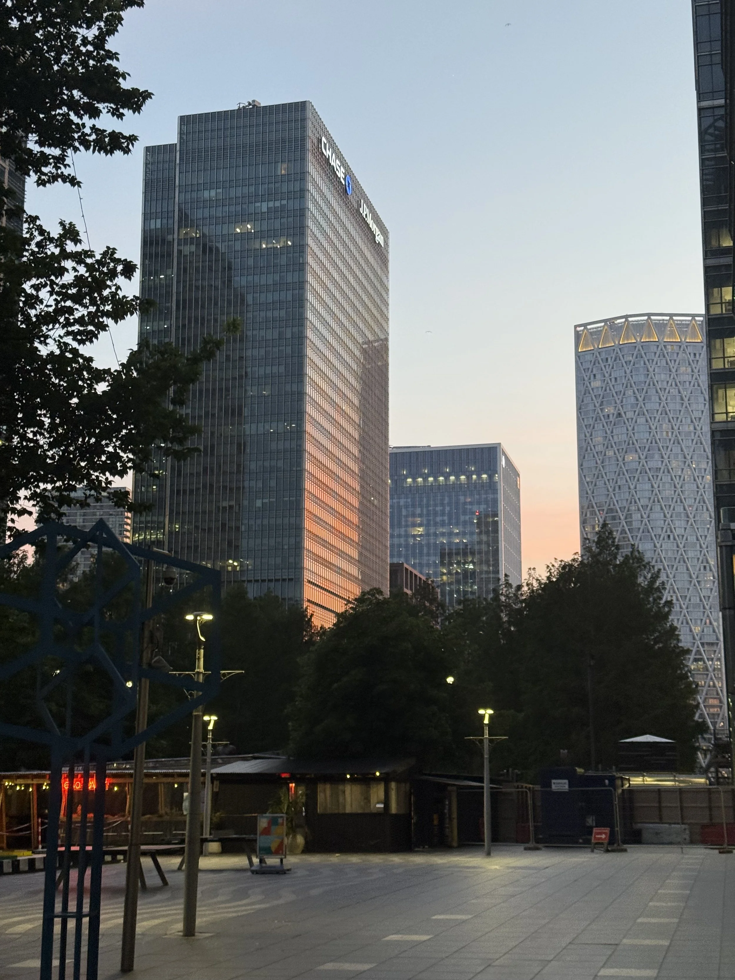 Tall office buildings in a city at dusk, with trees and a paved public area in the foreground.