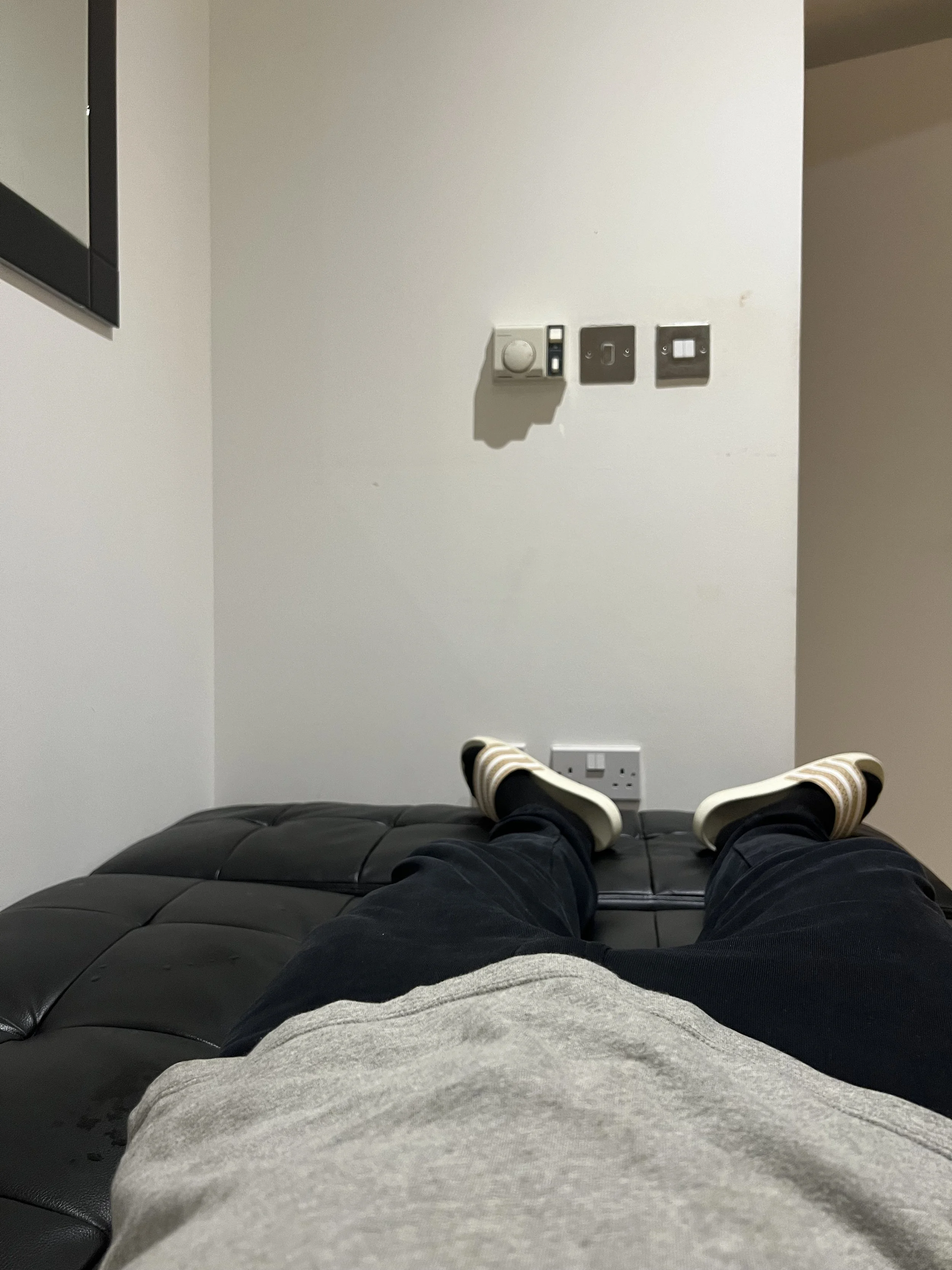 Person lying on a black ottoman with legs stretched out, wearing gray pants and striped slip-on shoes. The wall in front has several electrical switches, outlets, and a thermostat. Part of a chair is visible at the bottom edge of the image.