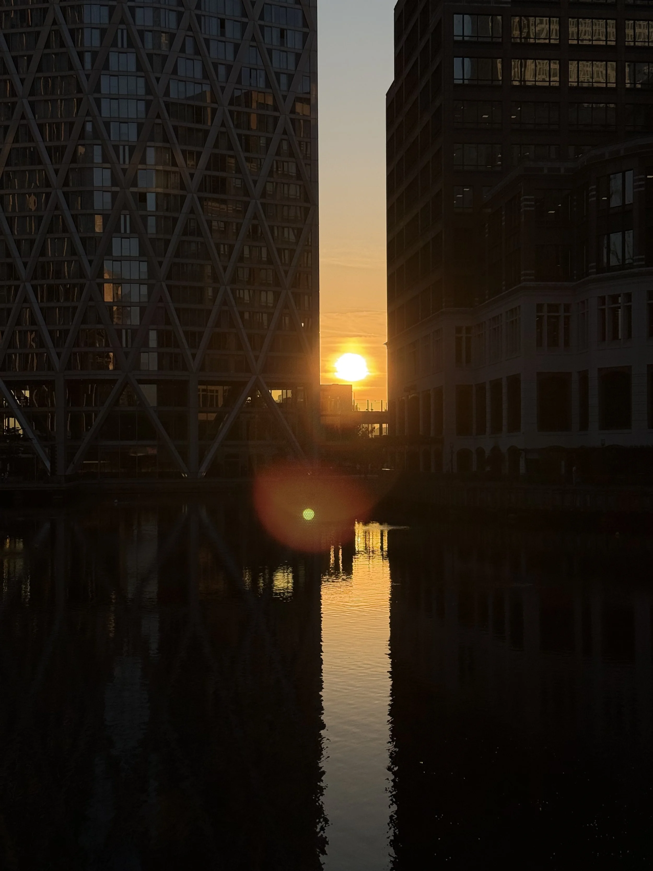Sunset behind modern high-rise buildings with glass facades reflecting the sunset colors, visible over a water body in an urban setting.