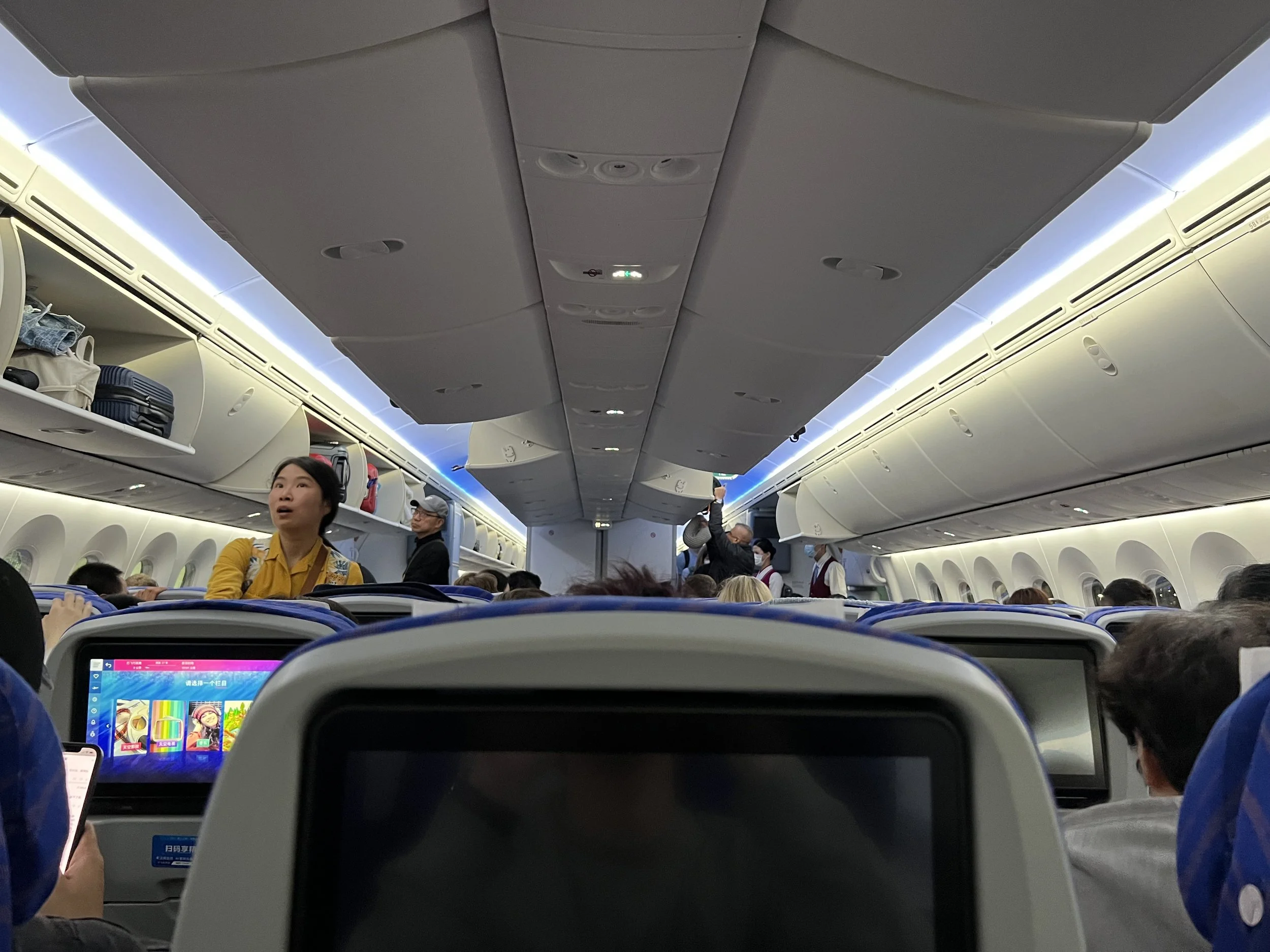 Inside view of an airplane cabin showing passengers seated, with overhead storage compartments and a woman standing by her seat and other travelers around her.
