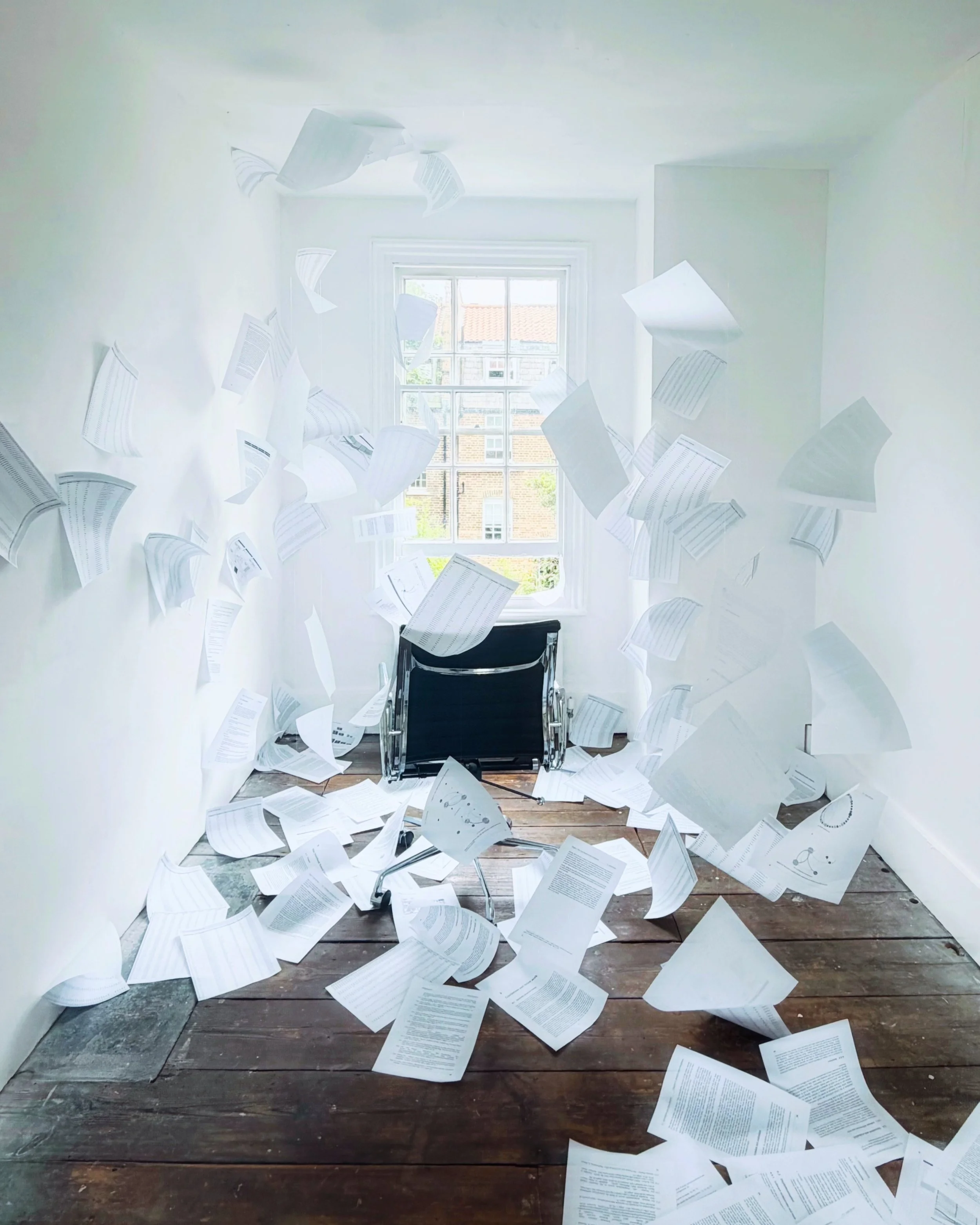 A room with scattered papers flying from the ceiling and floor. In the center, there is an empty wheel chair with black backrest and armrests, facing a large white window with multiple panes, showing older brick buildings and rooftops outside.