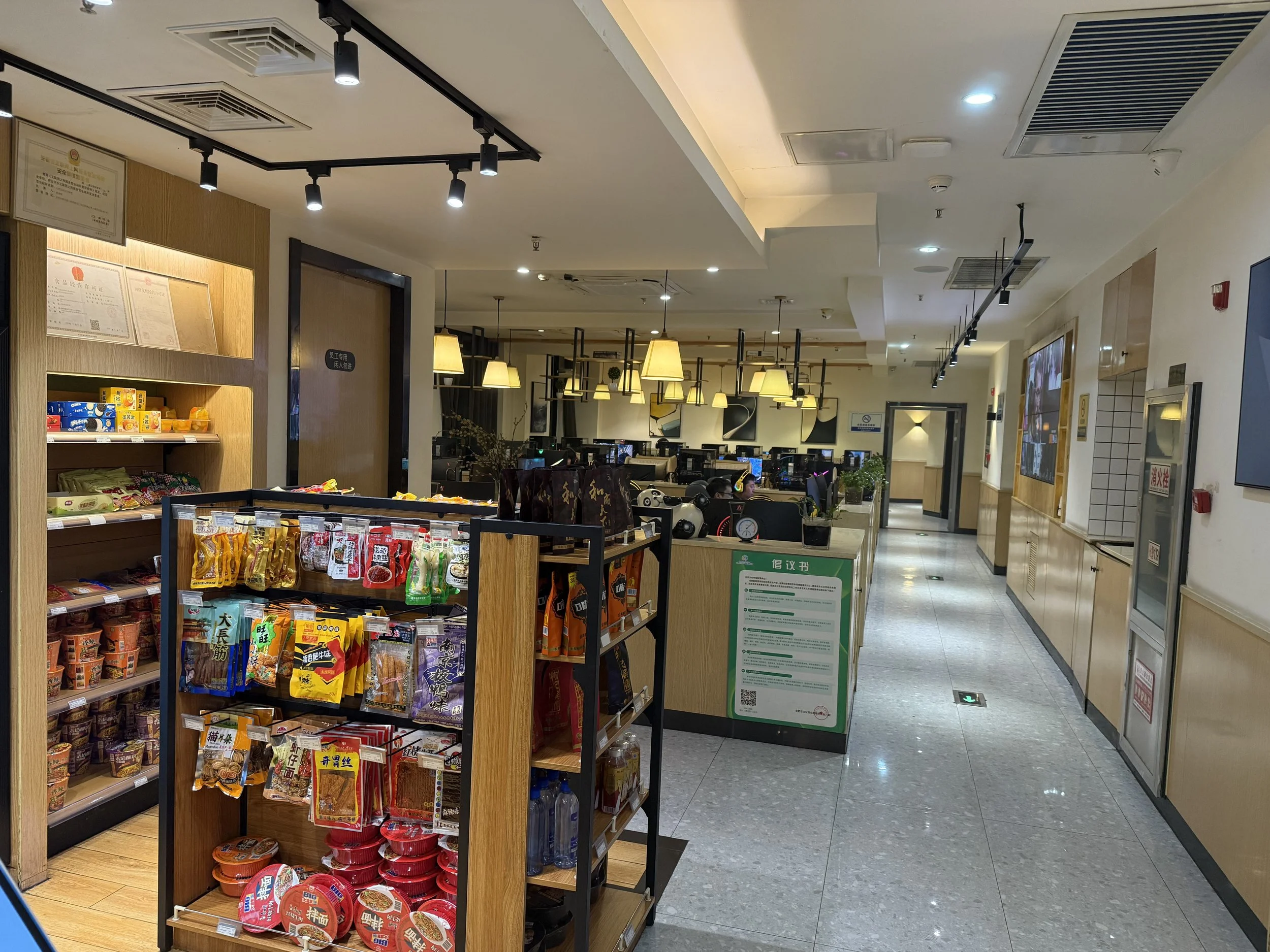 Inside a supermarket or convenience store with shelves stocked with snacks, drinks, and instant noodles in the foreground, and a seating area with tables and bright hanging lamps in the background.