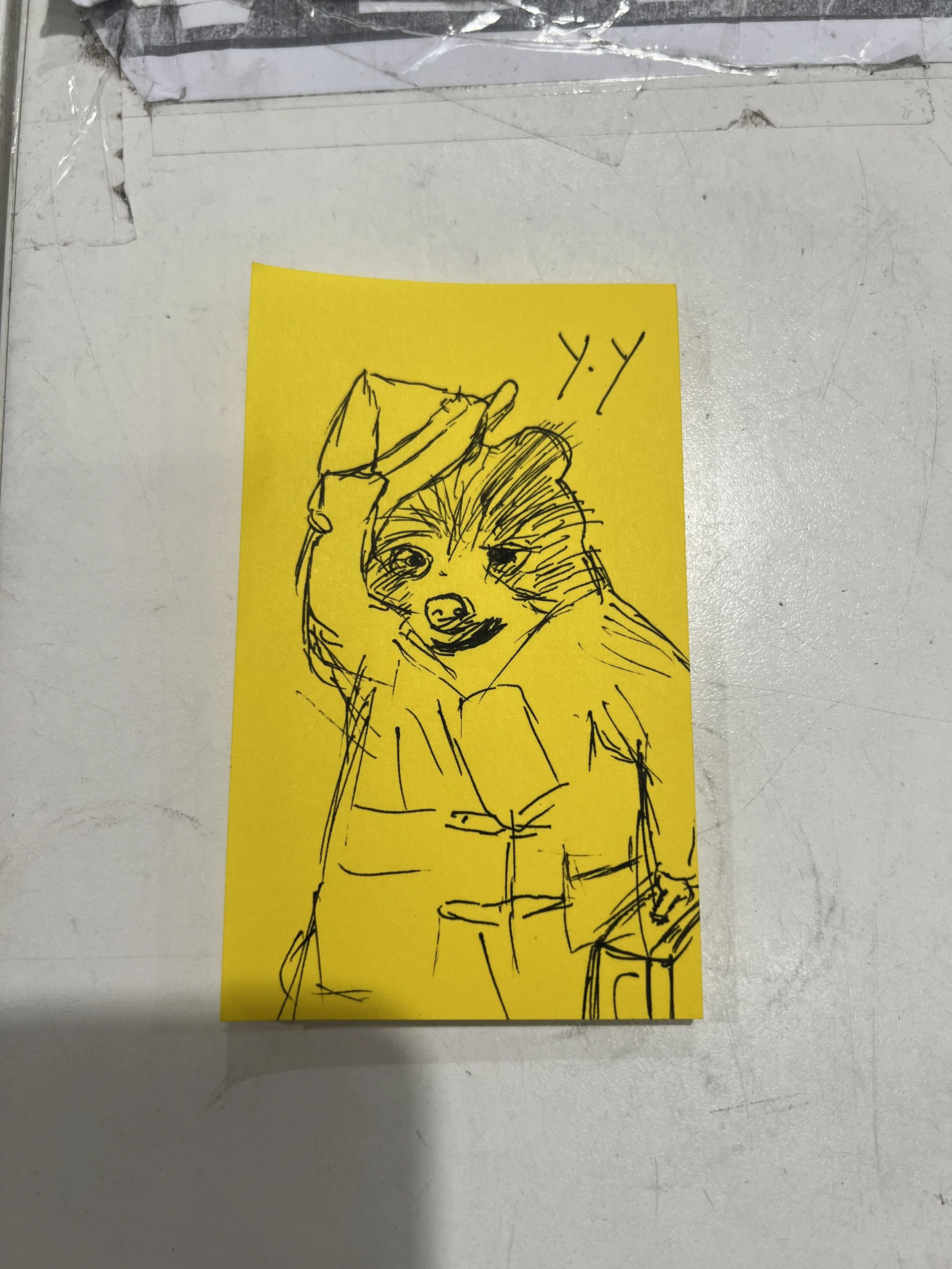 Simple sketch of a person with long hair, holding a book or paper and looking out from the side, drawn in black ink on a yellow sticky note.