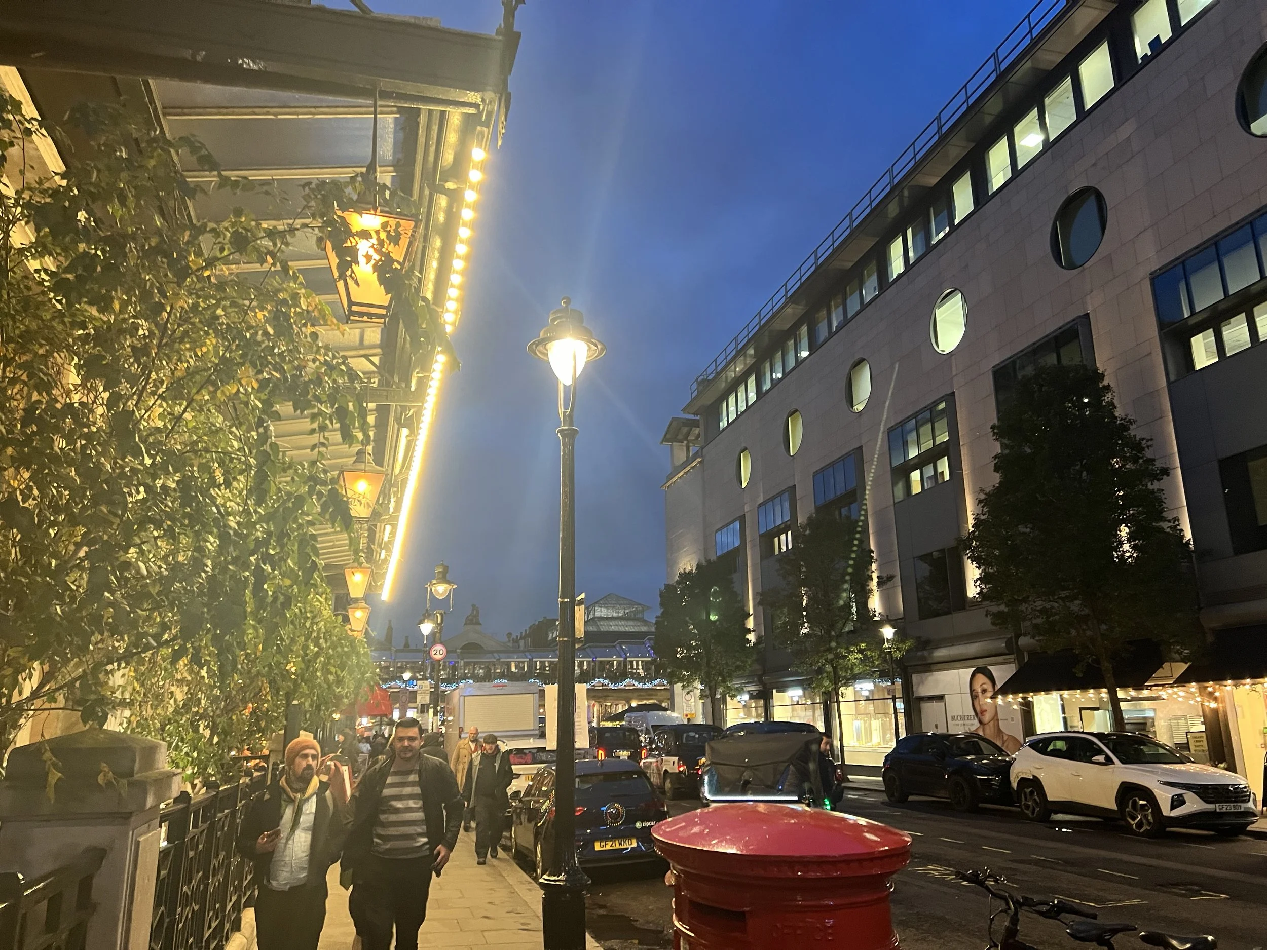 People walking along a city street at dusk, with illuminated buildings and streetlights, parked cars, and outdoor café seating visible.