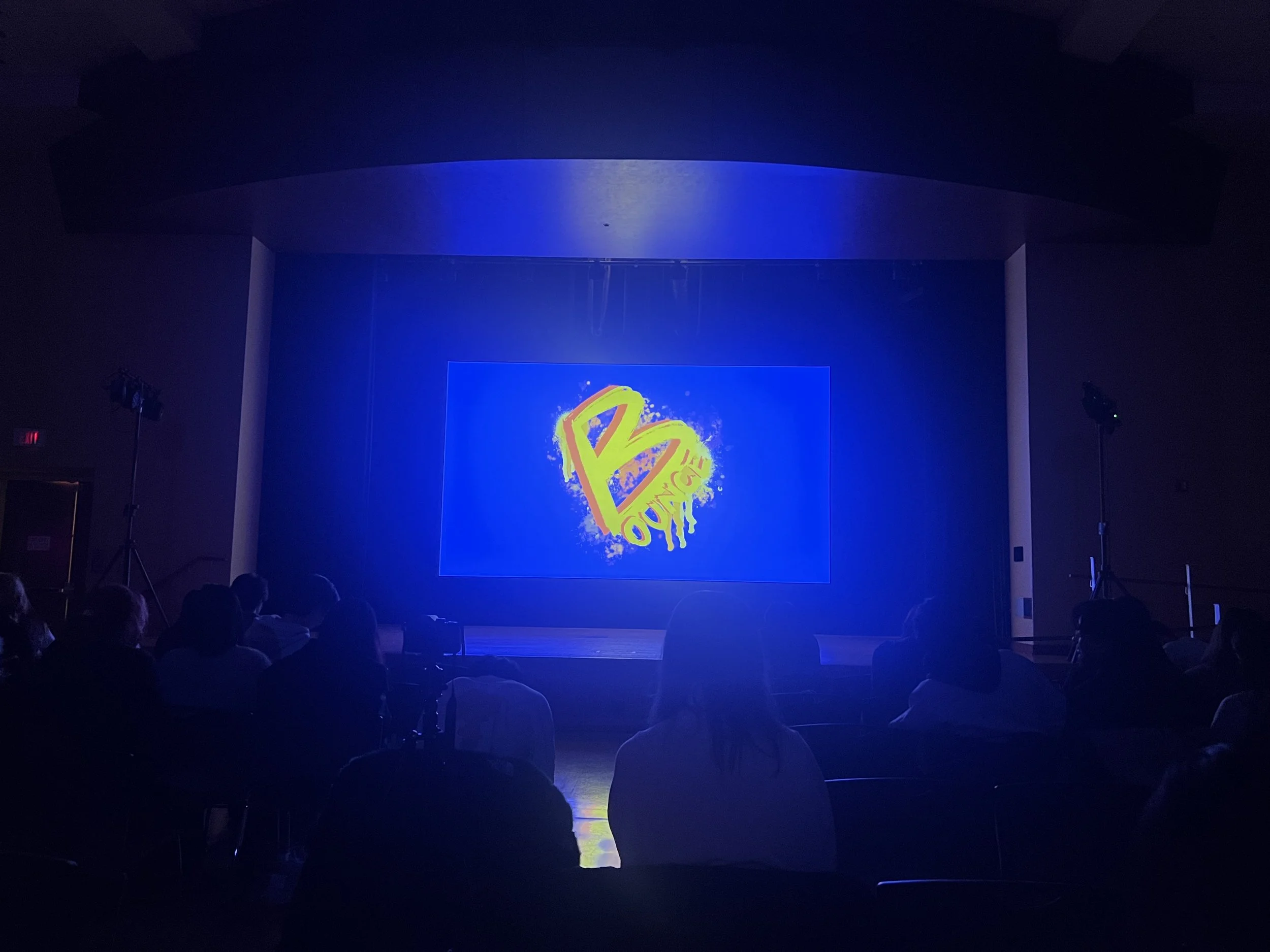 Dark auditorium with a stage illuminated by blue and purple lights, a large screen displaying colorful graffiti-style artwork, and audience members seated in the foreground.