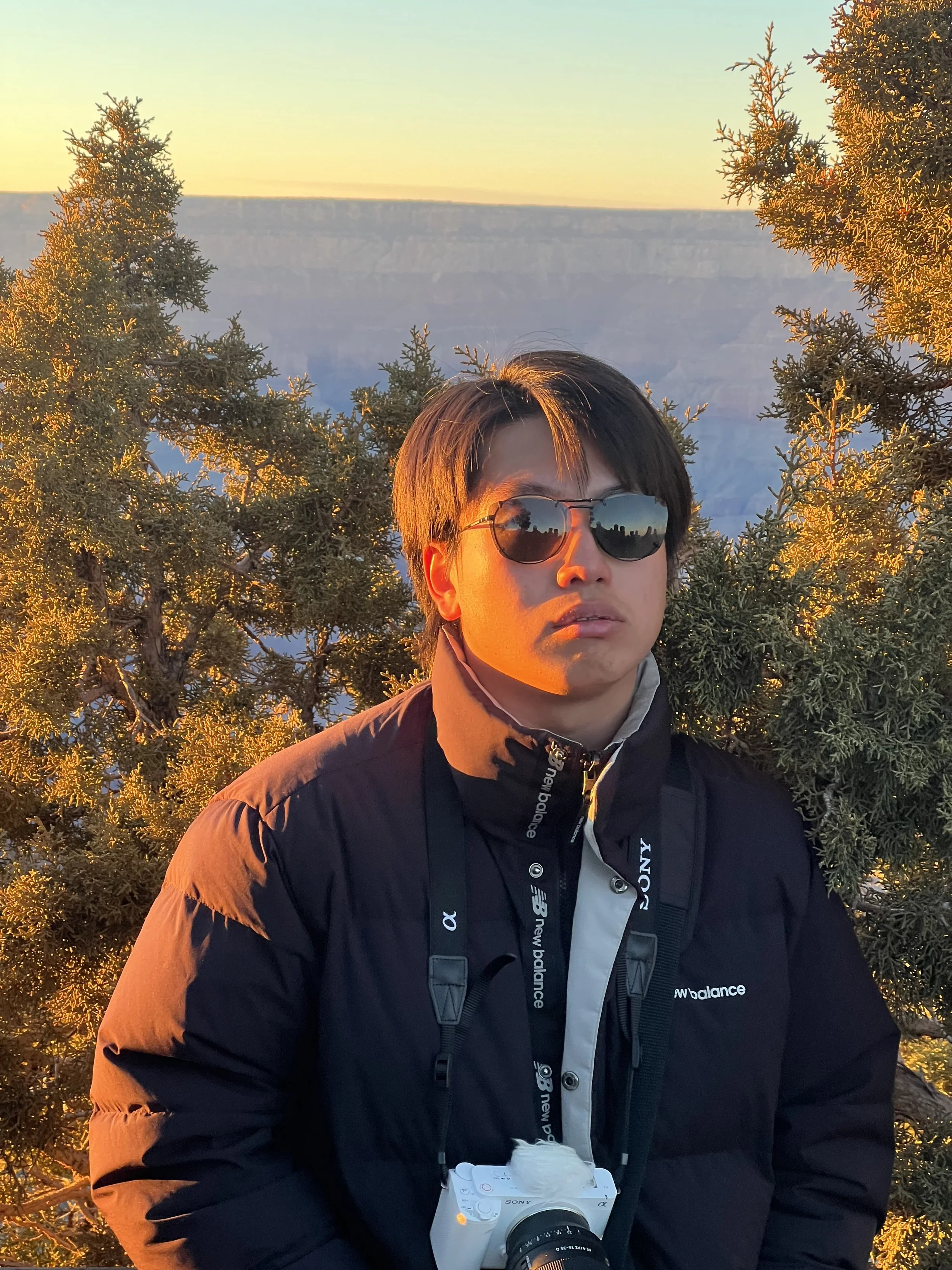 A young man with short black hair wearing sunglasses, a black jacket, and a camera strap around his neck, standing outdoors during sunset with a view of canyon cliffs in the background, surrounded by pine trees.
