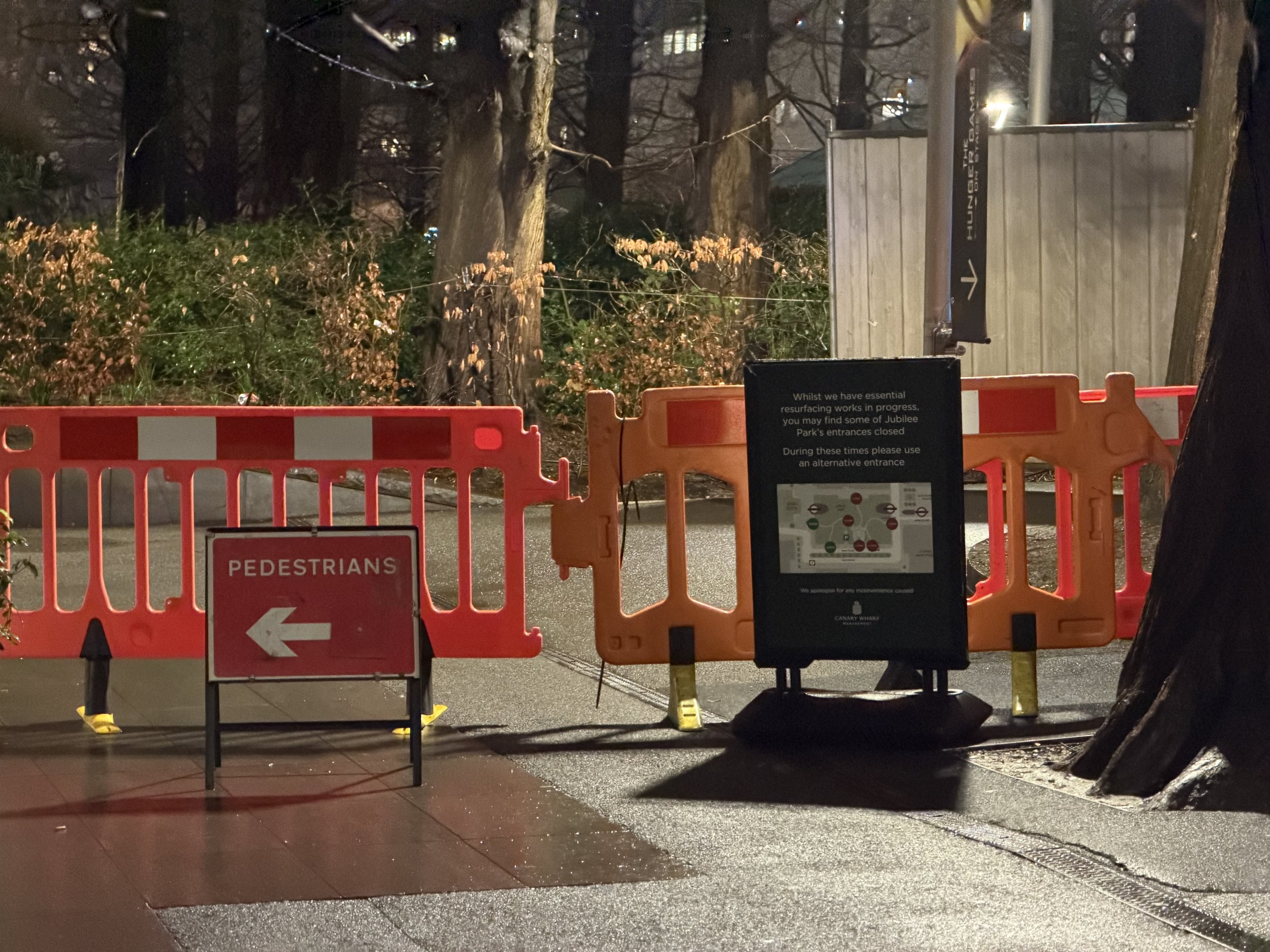 Nighttime view of a street blocked by orange construction barriers and signs directing pedestrians and vehicles. Behind the barriers, there are leafless trees and a wooden structure with a sign indicating a different entrance. The scene is illuminate