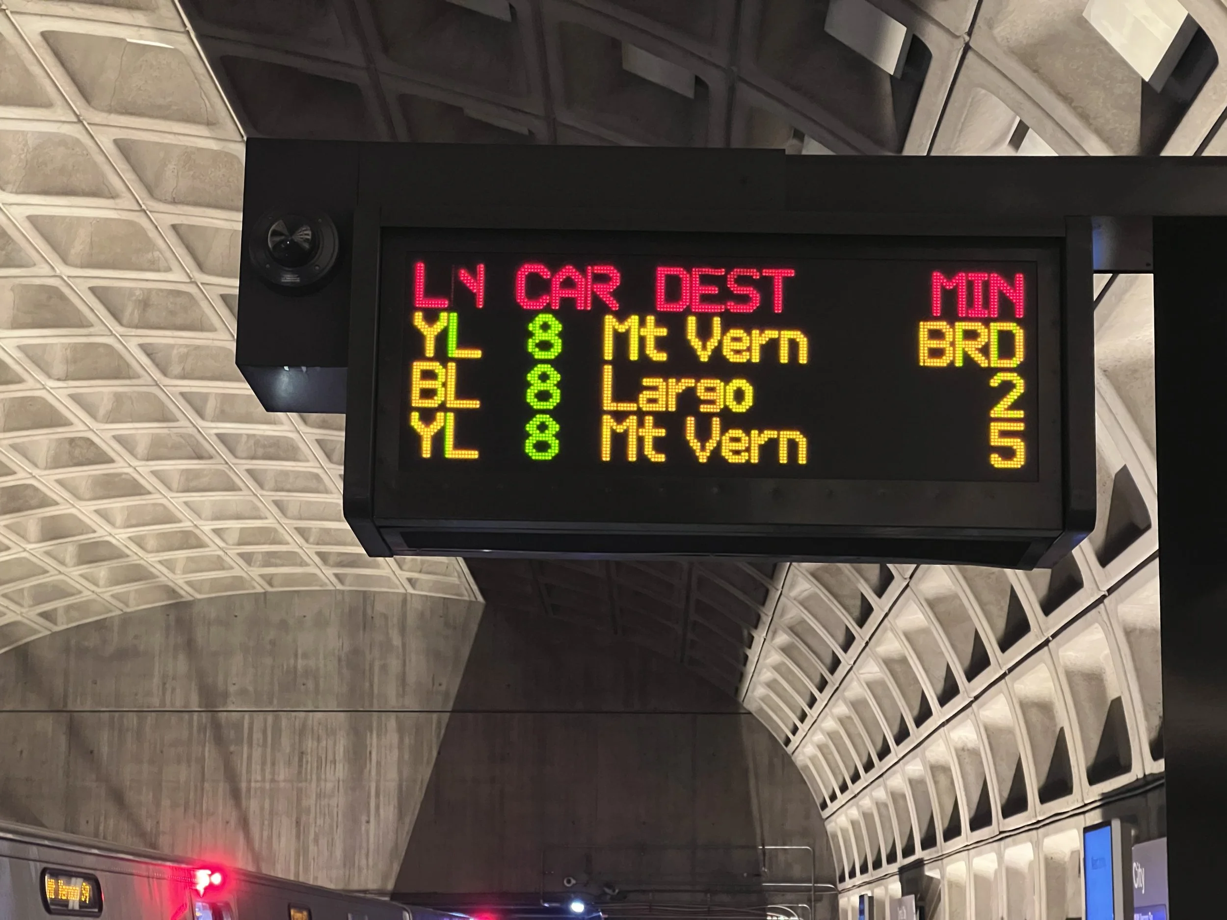 Electronic flight information display showing departure times and destinations at an airport, with ceiling and wall patterns in the background.