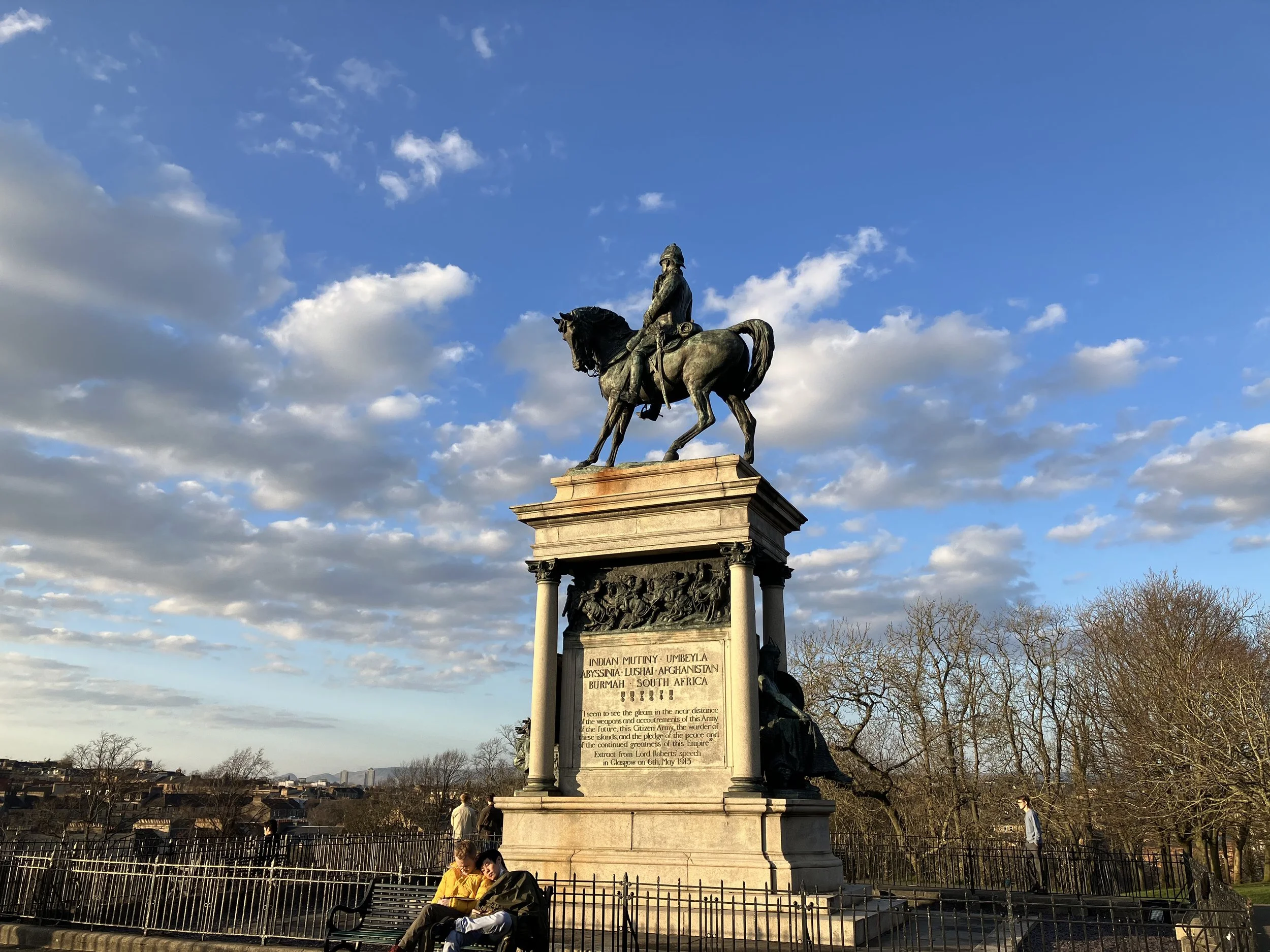 A bronze statue of a mounted warrior on a tall stone pedestal with inscriptions, set against a partly cloudy sky. People are sitting and walking nearby in a park.