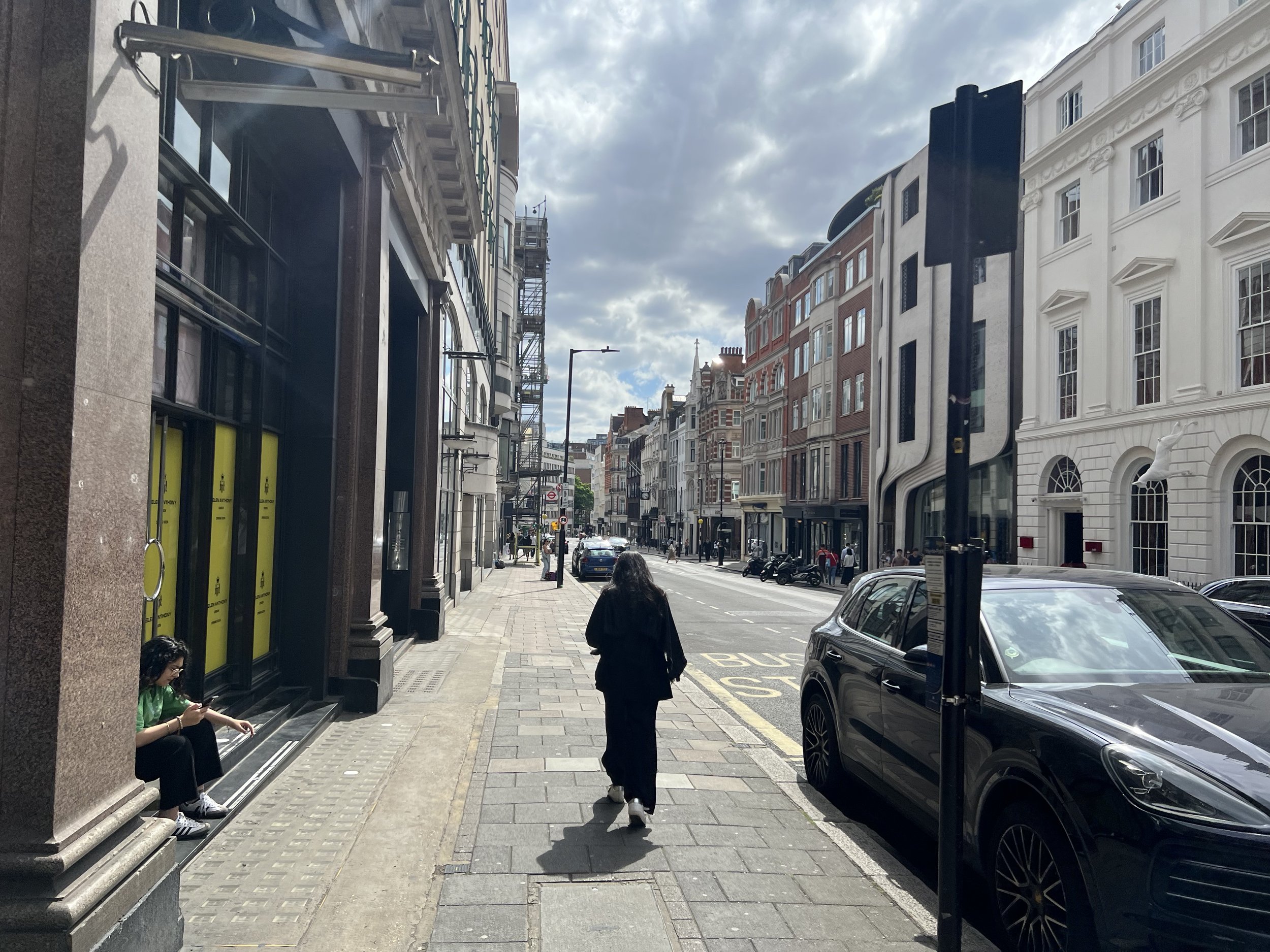 City street scene with people walking on the sidewalk, parked cars, and multi-story buildings with ornate facades and balconies. The sky is partly cloudy.