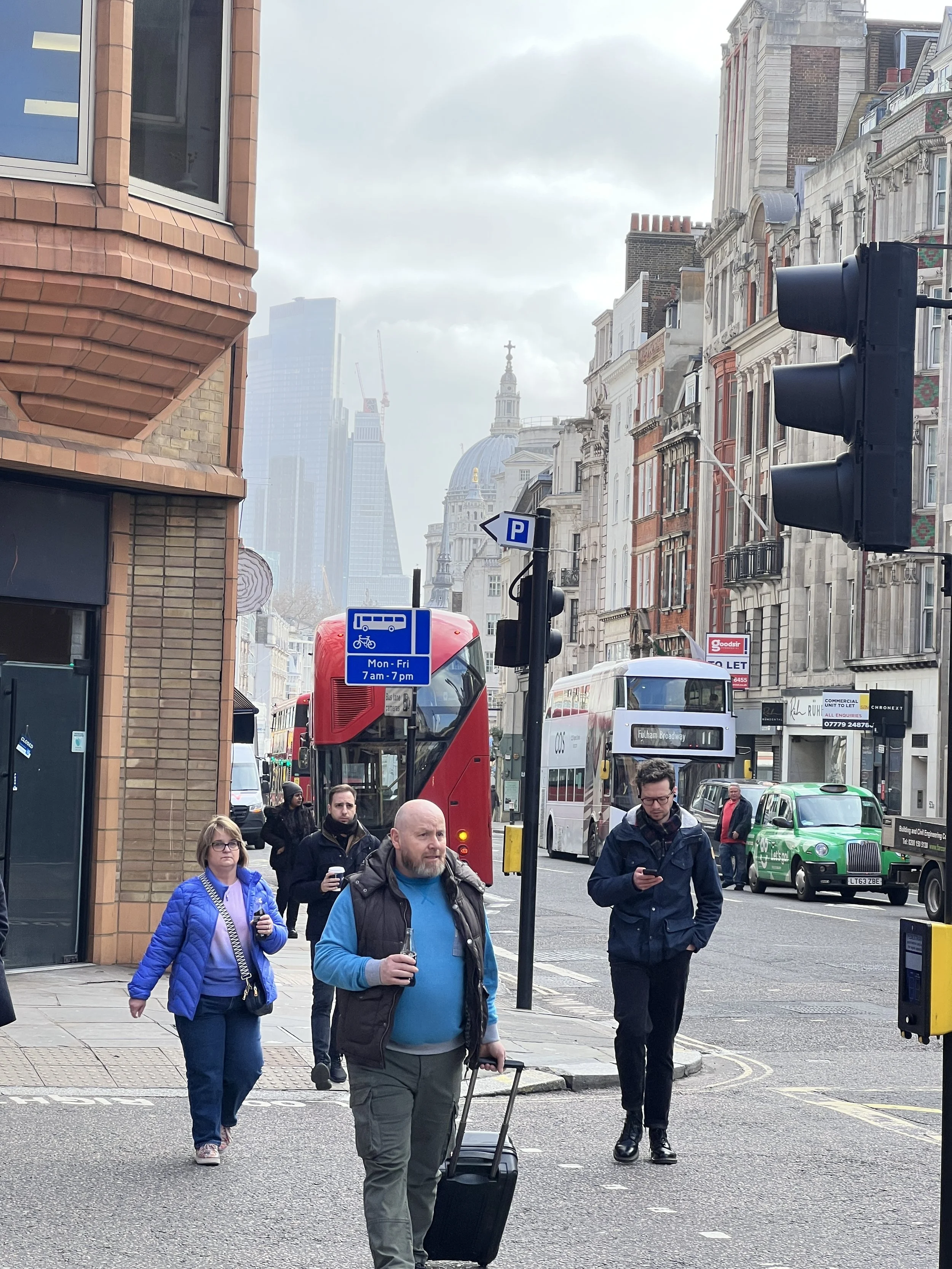 People walking on a city street with buses, cars, and historic and modern buildings, including a domed structure with a cross on top, under cloudy skies.