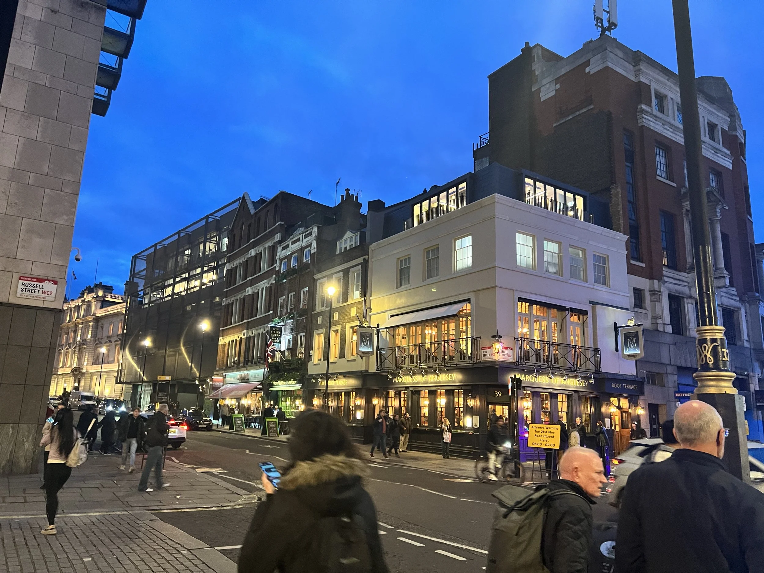 A street scene in the evening with people walking and cars parked along the road. Tall buildings with lit windows line the street, and a restaurant named 'Marquess of Anglesey' is visible on the ground level. The sky is dark blue, indicating twilight