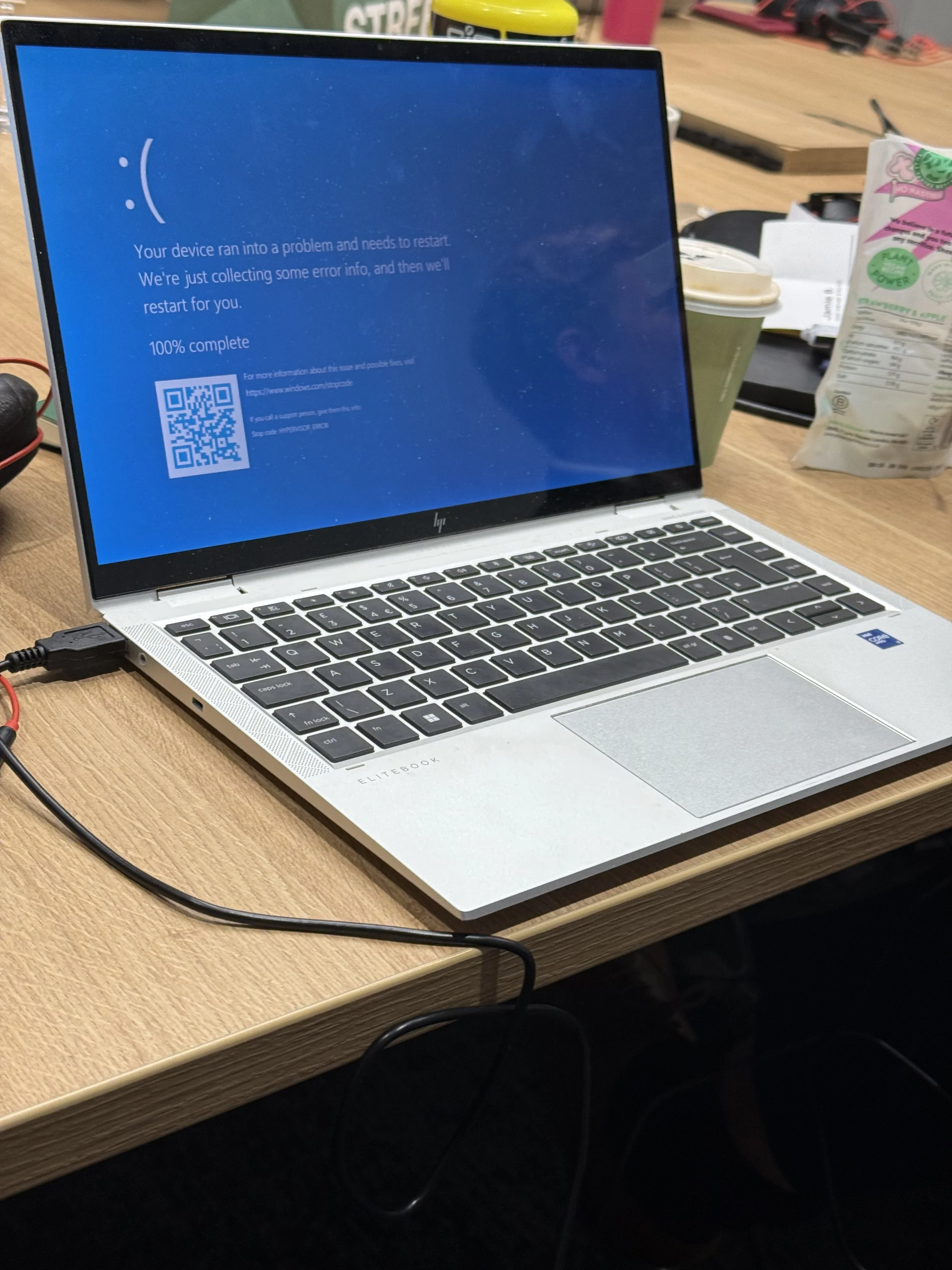 A laptop displaying a blue screen error message stating 'Your device ran into a problem and needs to restart,' with a QR code and instructions for more information, on a wooden desk with food and drink items surrounding it.